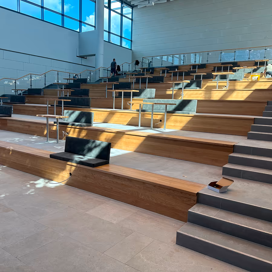 Modernes Auditorium mit hohen Fenstern, abgestuften Holzbänken mit kleinen Tischen und einzelnen Sitzpolstern.