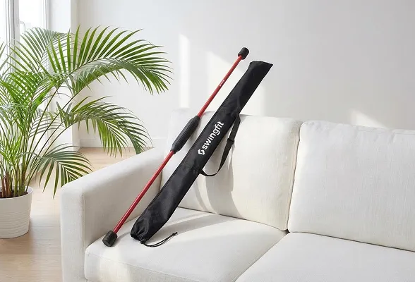 Red golf training stick leaning against a black Swingfit bag on a white sofa with a potted plant nearby.