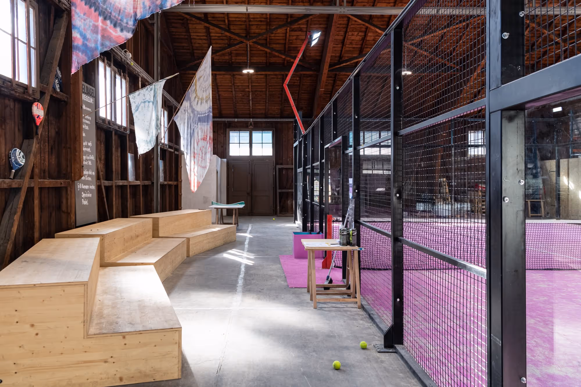 Innenansicht einer Halle mit Holzbänken, bunten Stoffen an der Wand und einem abgetrennten Bereich mit pinkem Bodenbelag und schwarzen Metallgittern.