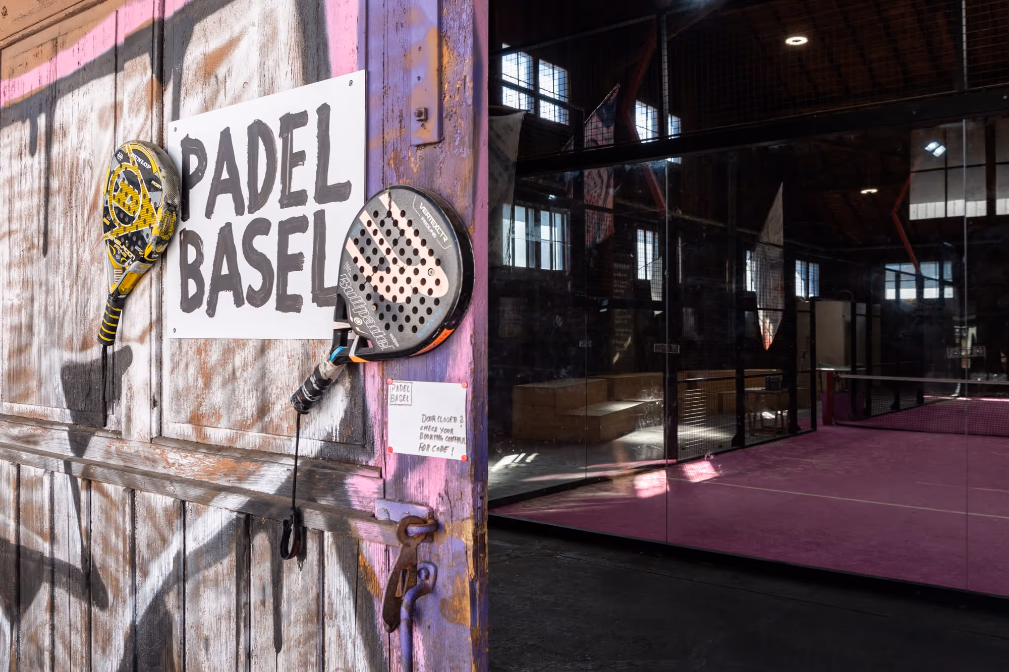Hell erleuchteter Padel-Tennisplatz mit pinkem Boden und zwei Padel-Schlägern neben einem Schild mit der Aufschrift 'Padel Basel' an einer bemalten Holztür.