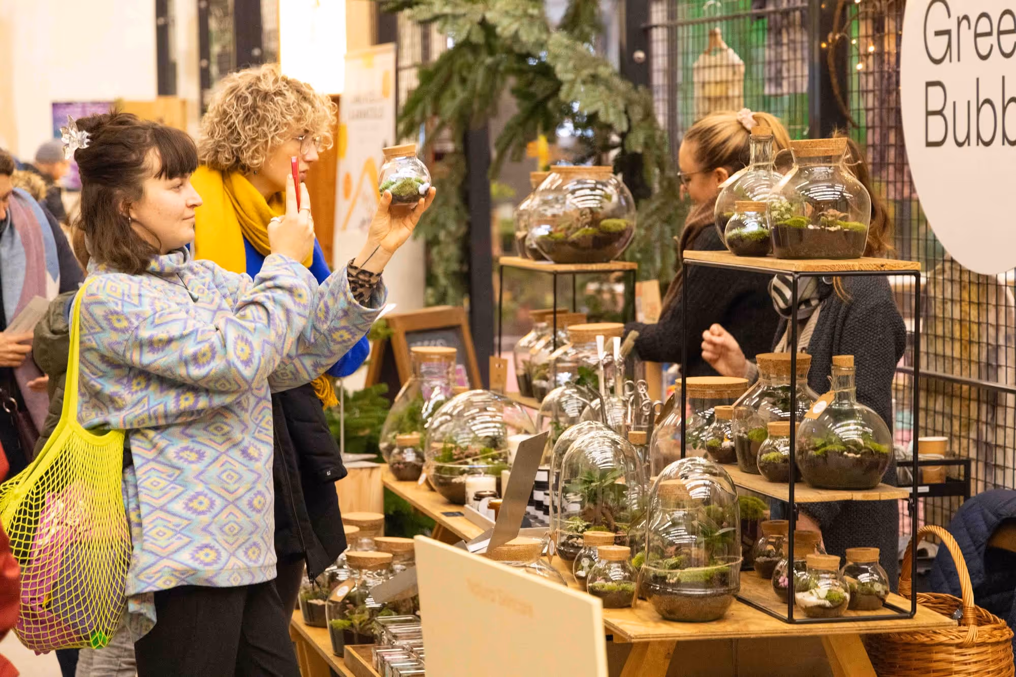 Personen betrachten und fotografieren Glasgefässe mit Pflanzen und Moos an einem Marktstand mit Holzregalen.