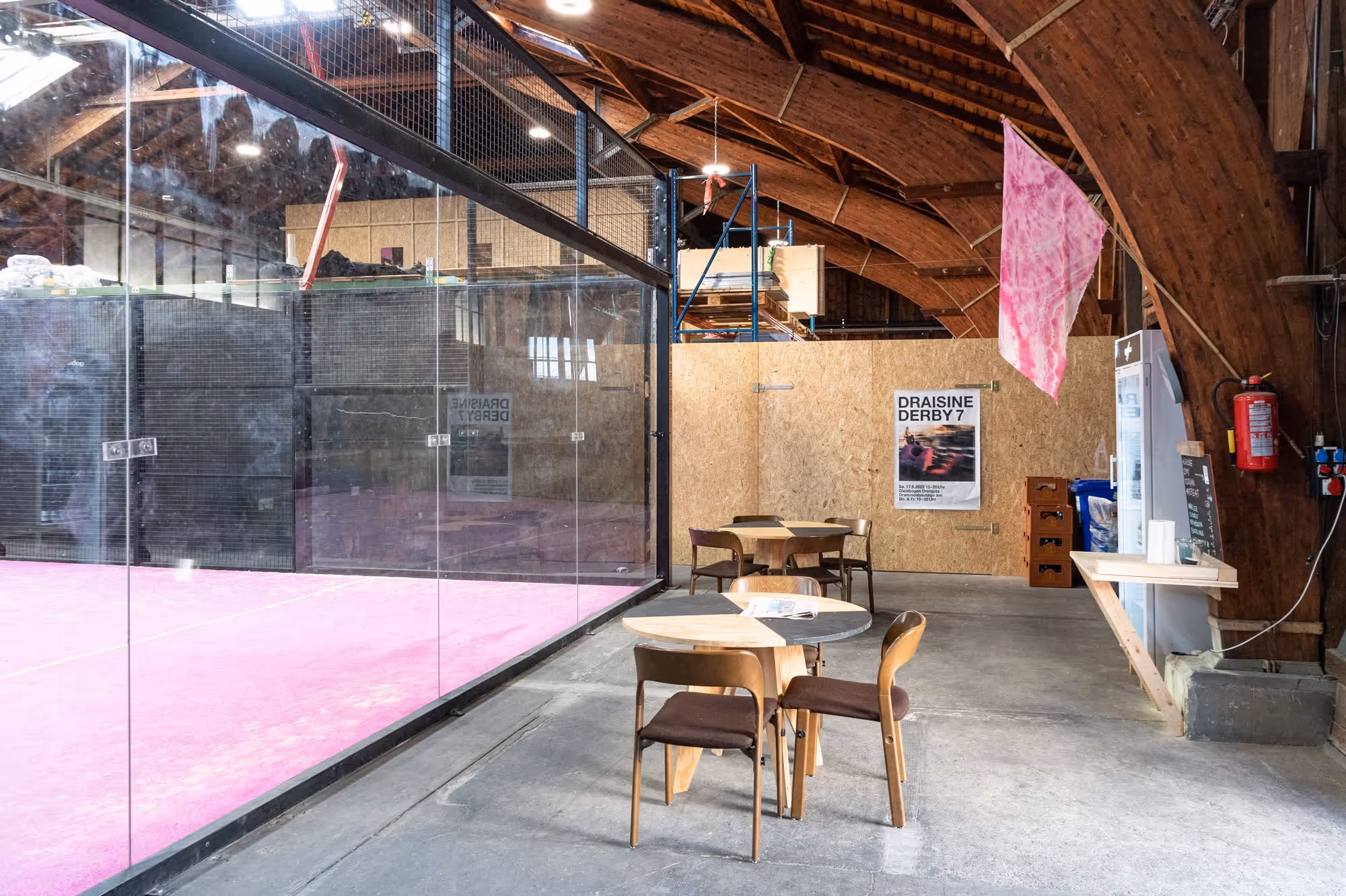 Innenbereich mit pinkfarbenem Squash- oder Padelplatz hinter Glas, davor Tische und Stühle zum Sitzen in einer Halle mit Holzdecke.