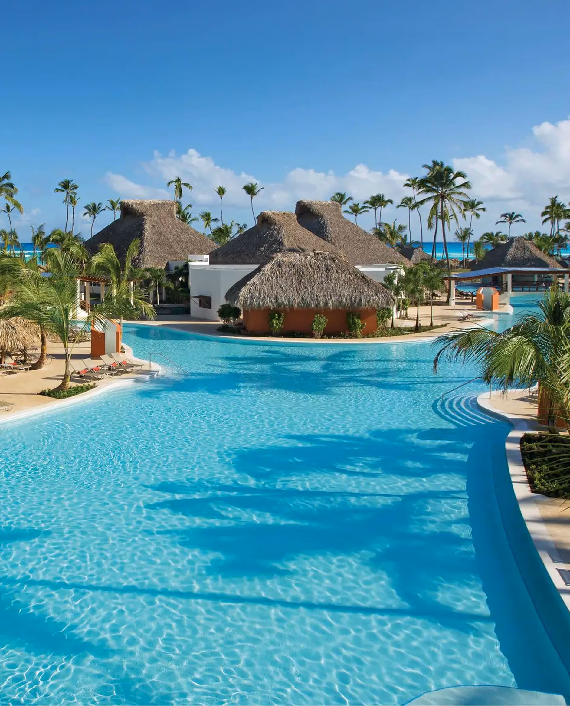 Large blue pool with palm trees and thatched roof huts under clear blue sky at seaside resort.