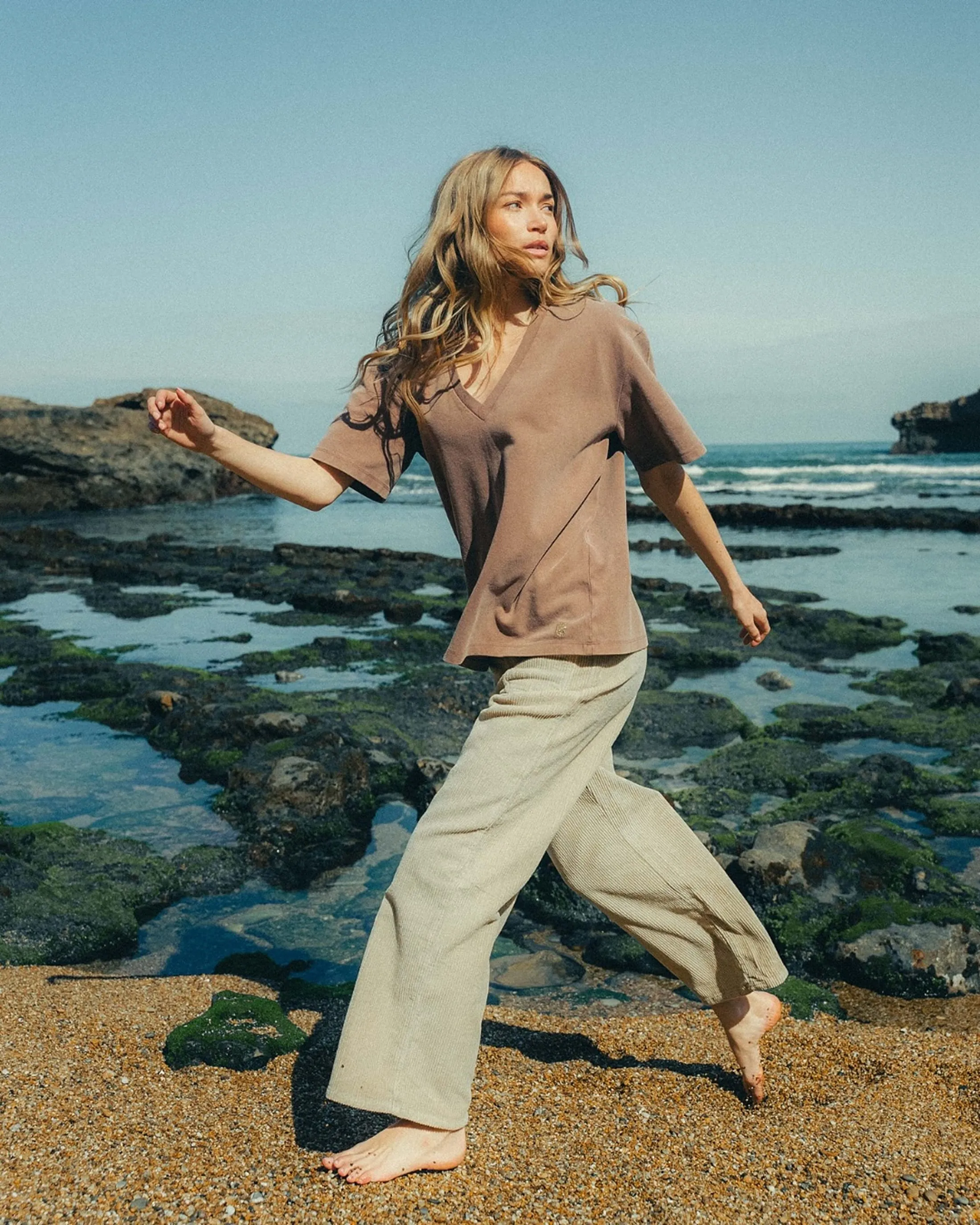 Woman in beige pants and brown shirt walking barefoot on rocky beach near ocean waves.