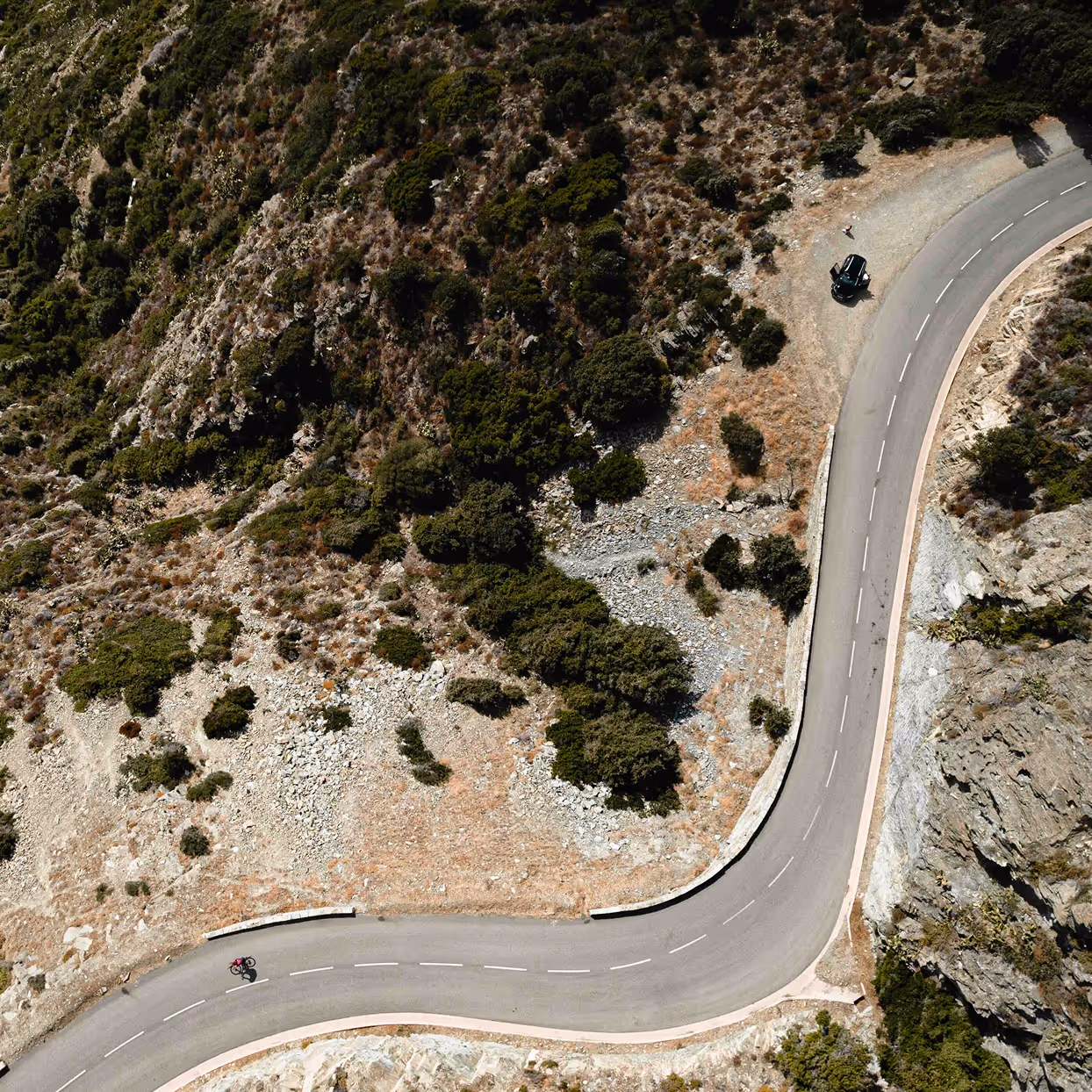 A cyclist on a winding mountain road near a parked black car by rocky terrain and bushes.