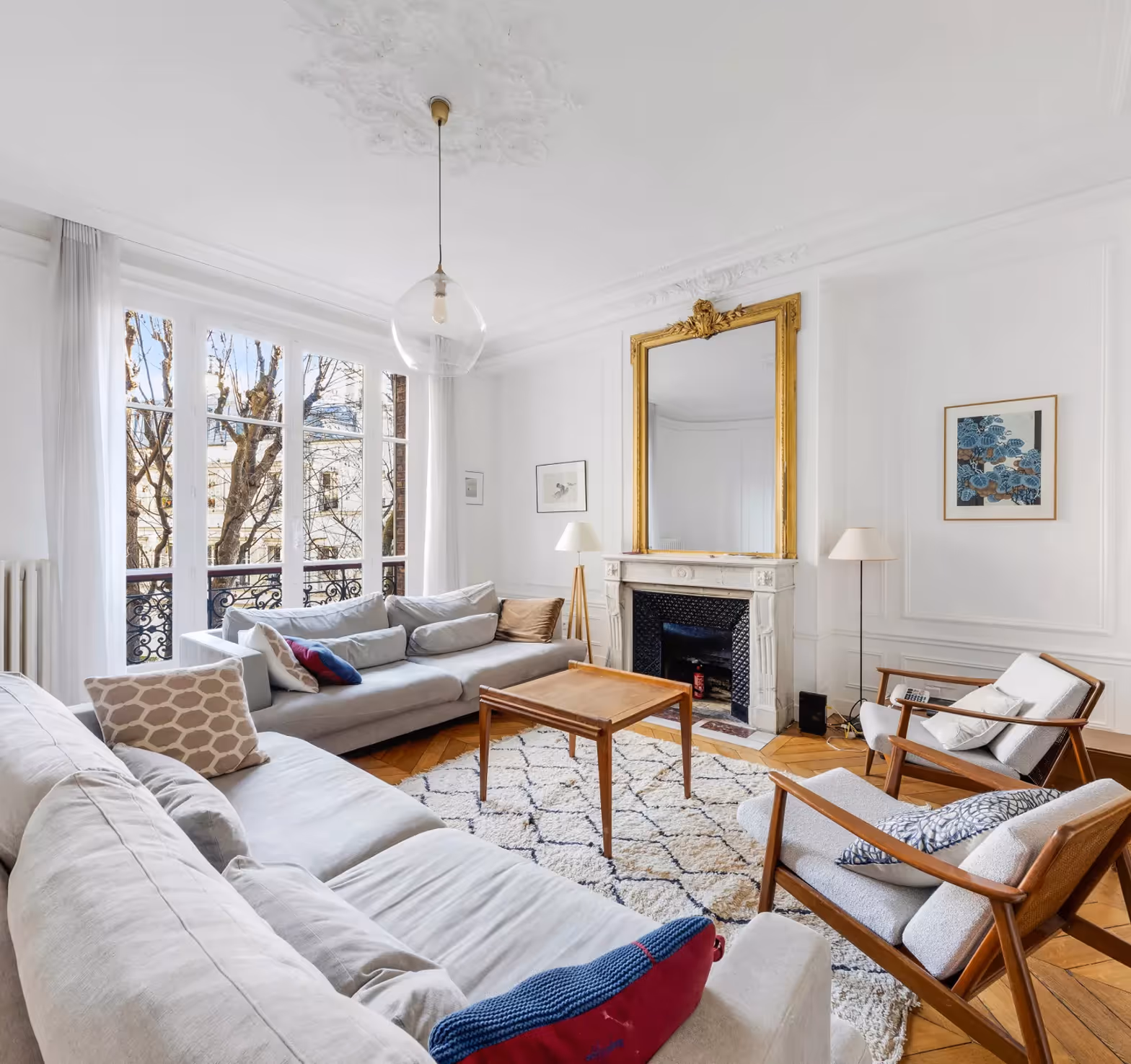 Bright living room with large windows, white sofas, wooden chairs, a marble fireplace, and a wooden coffee table.