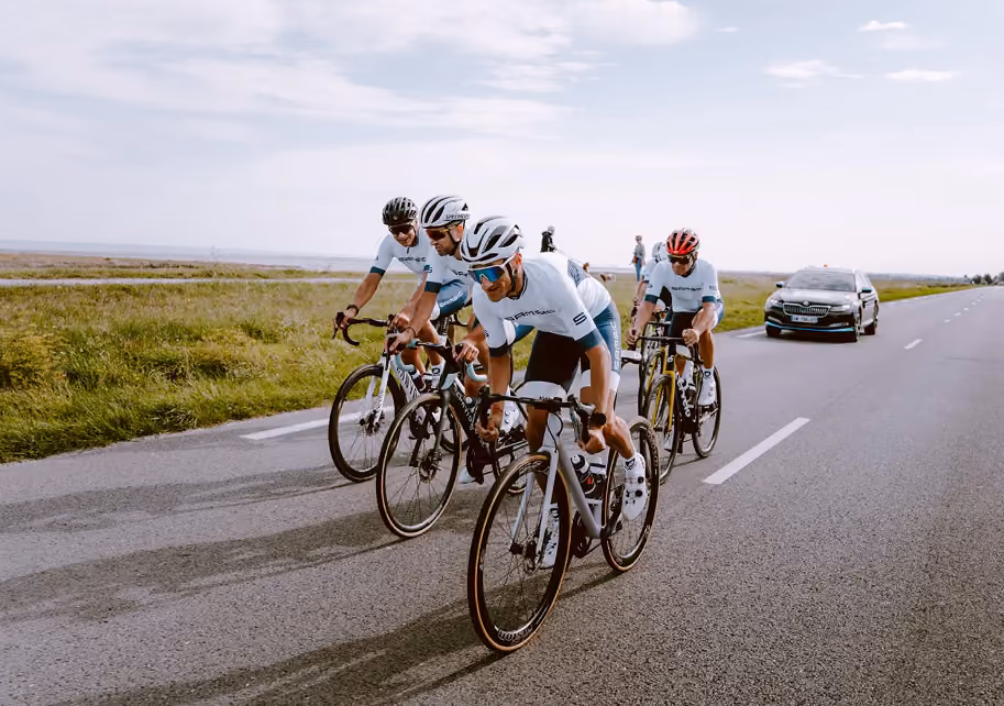 Four cyclists in matching gear riding on a road with grass fields and a car behind them.