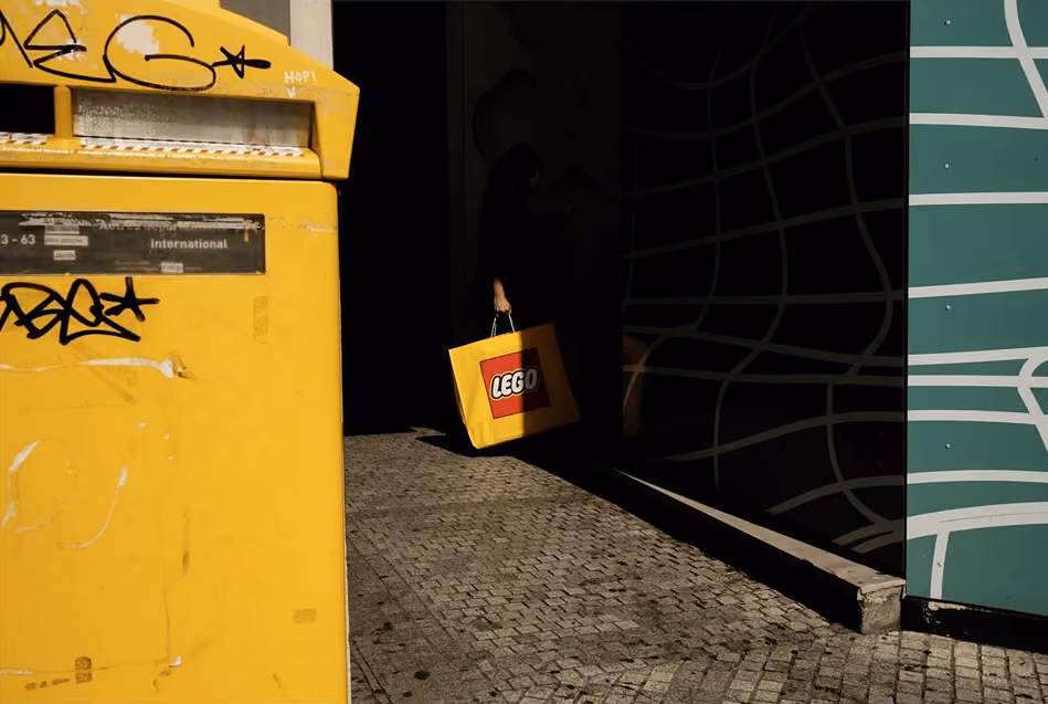 Person carrying yellow LEGO shopping bag walking on a cobblestone path beside a yellow mailbox.