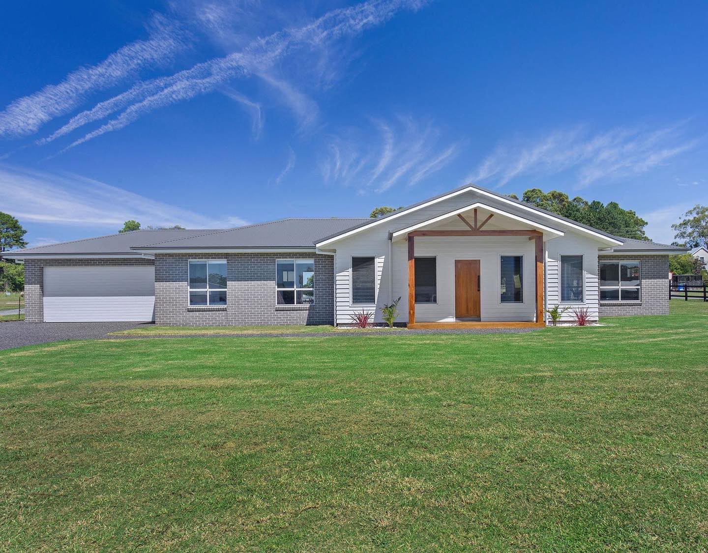 Single-story house with gray brick, white siding, wooden front door, and large green lawn under blue sky.