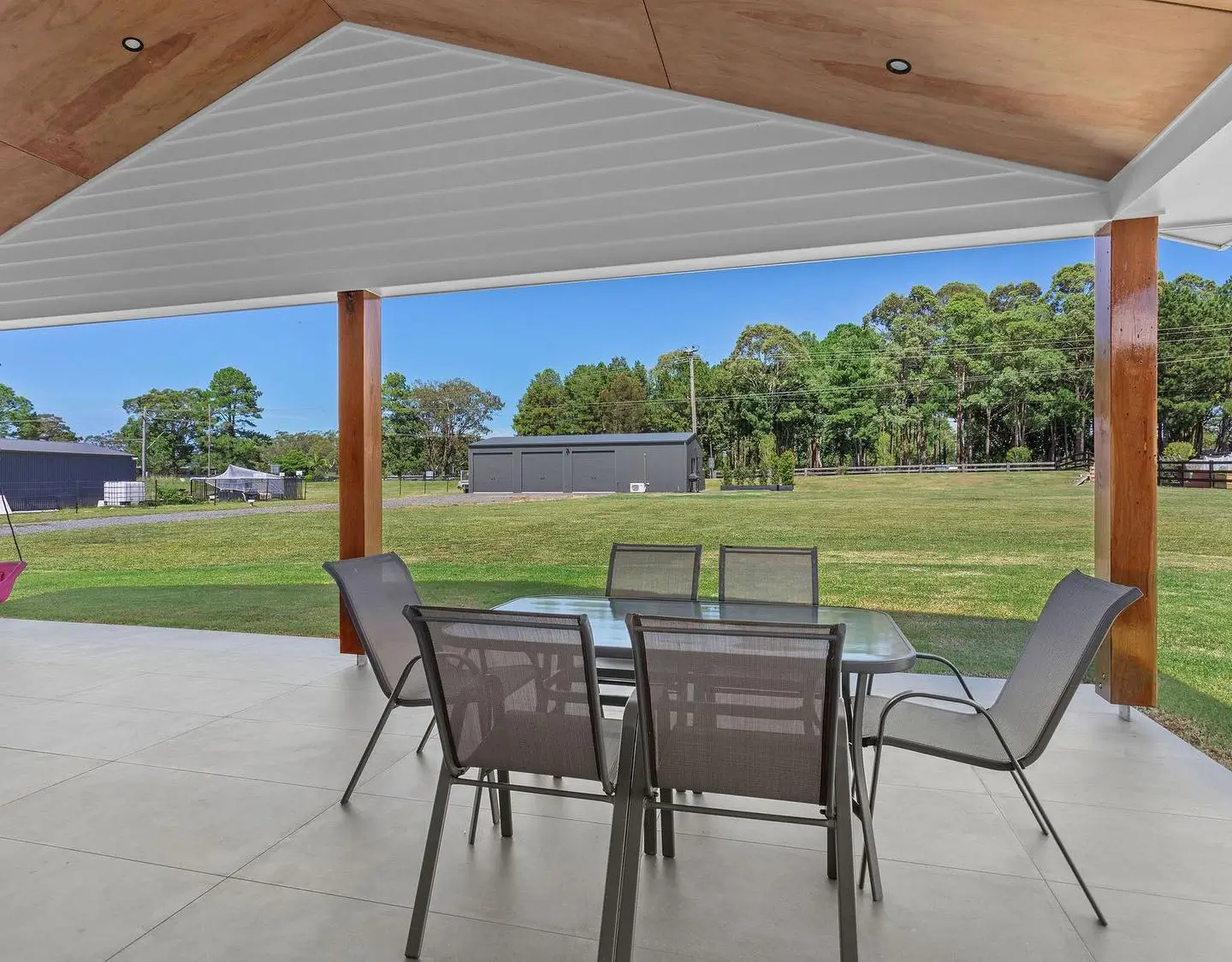 Outdoor covered patio with glass table and six mesh chairs overlooking a large grassy yard and trees.