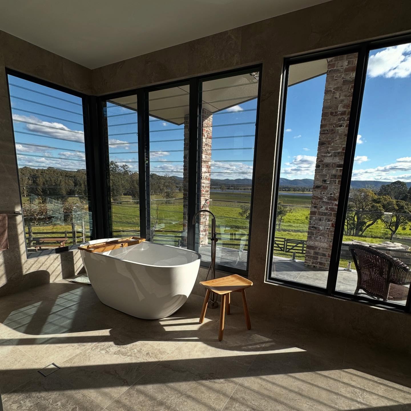 Modern bathroom with a white freestanding tub and large windows showing green fields and blue sky.