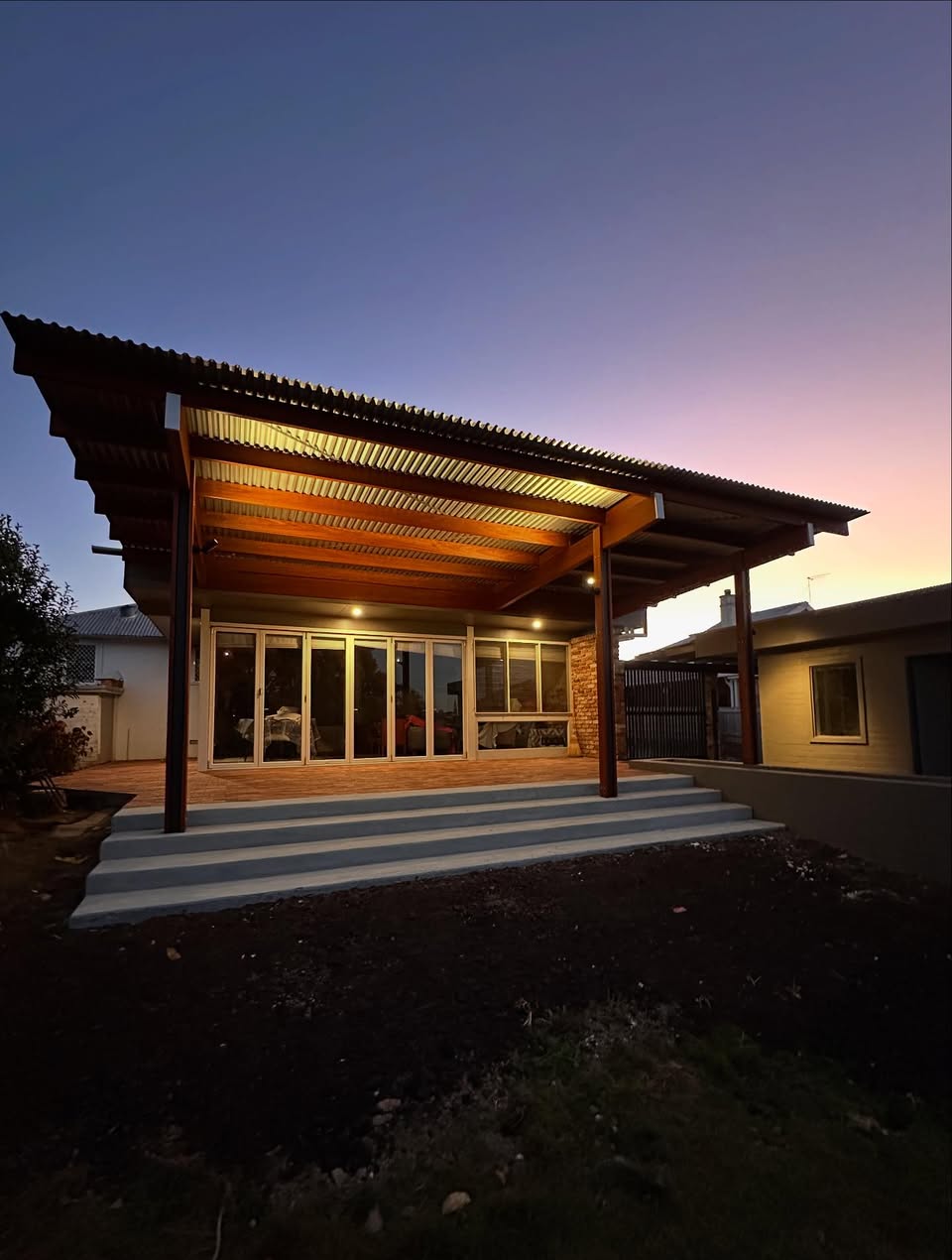 Back porch patio with wooden roof and steps at dusk, with house windows and soft lighting.