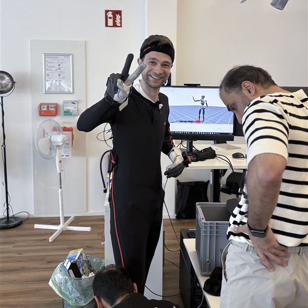 Behind the Scenes: lächelnde Person im Motion-Capture-Anzug zeigt ein Victory-Zeichen, während ein Kollege auf einen Bildschirm blickt