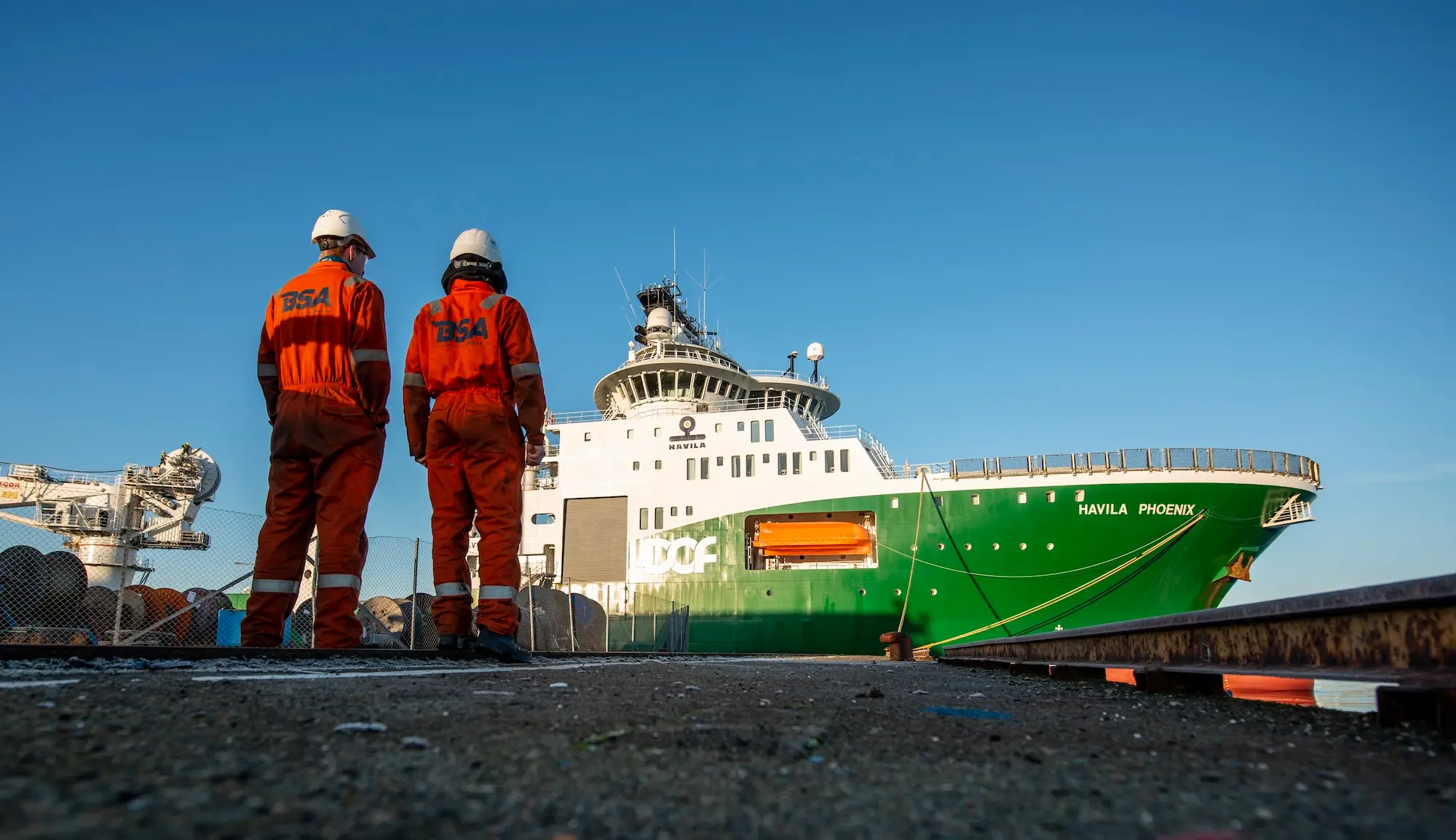 Two workers in orange coveralls and helmets stand on dock beside the green and white ship.