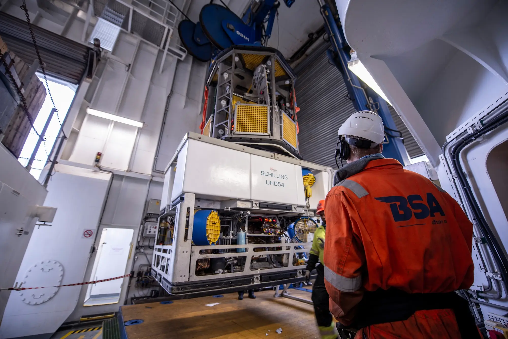 Worker in orange BSA uniform and helmet overseeing lifting of large industrial equipment indoors.