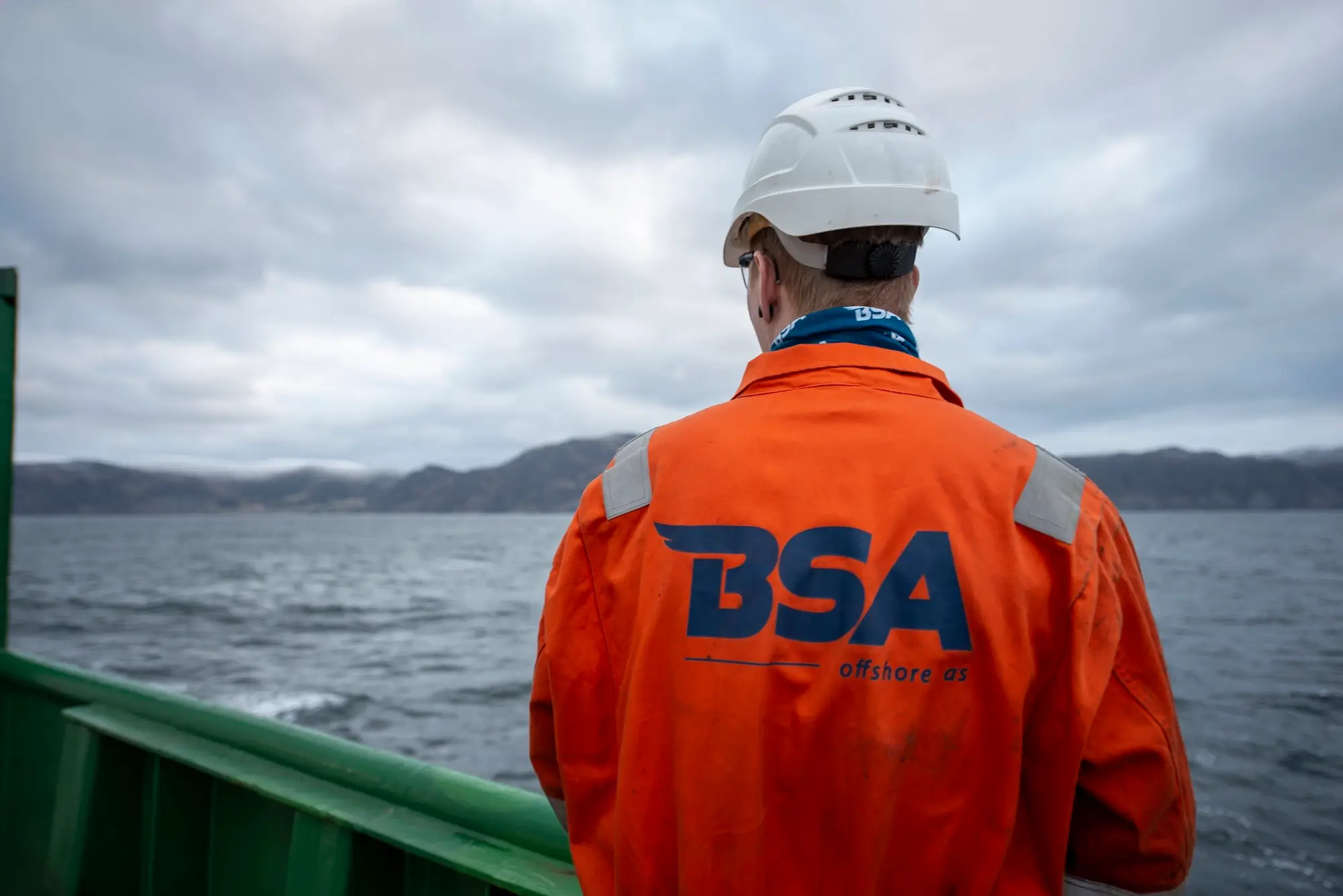 Person in orange BSA offshore jacket and white helmet on a boat facing the sea with mountains in background.