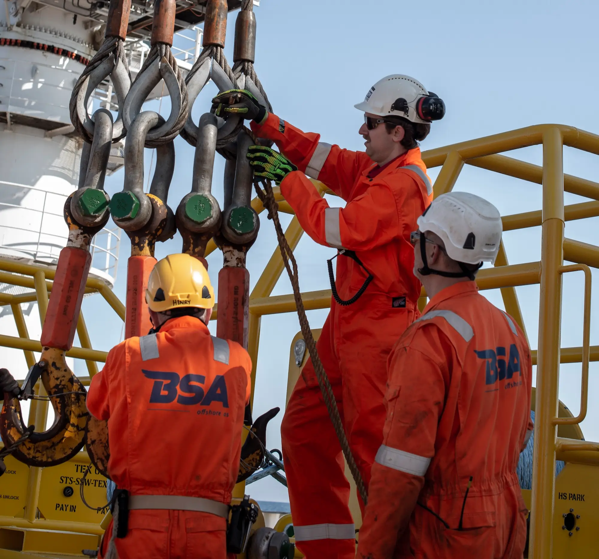 Three offshore workers in orange gear and helmets handling large metal hooks on a rig.
