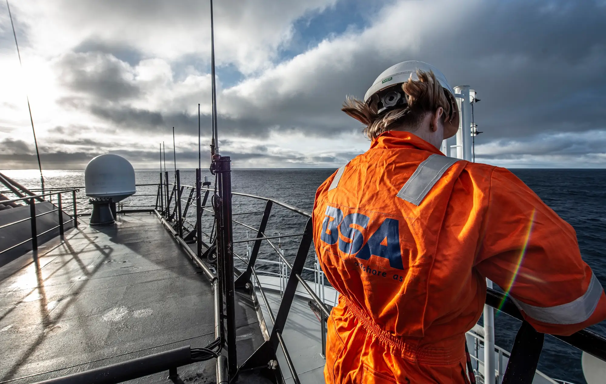 Worker in orange safety gear and helmet standing on ship deck overlooking the ocean under cloudy sky.