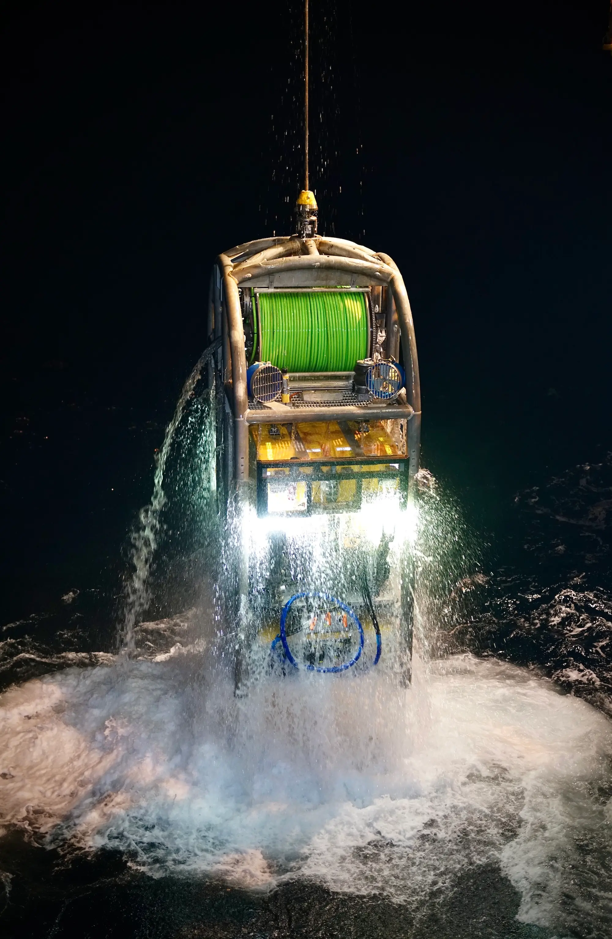 Underwater remotely operated vehicle with bright lights and green cable spool, emerging from water at night.