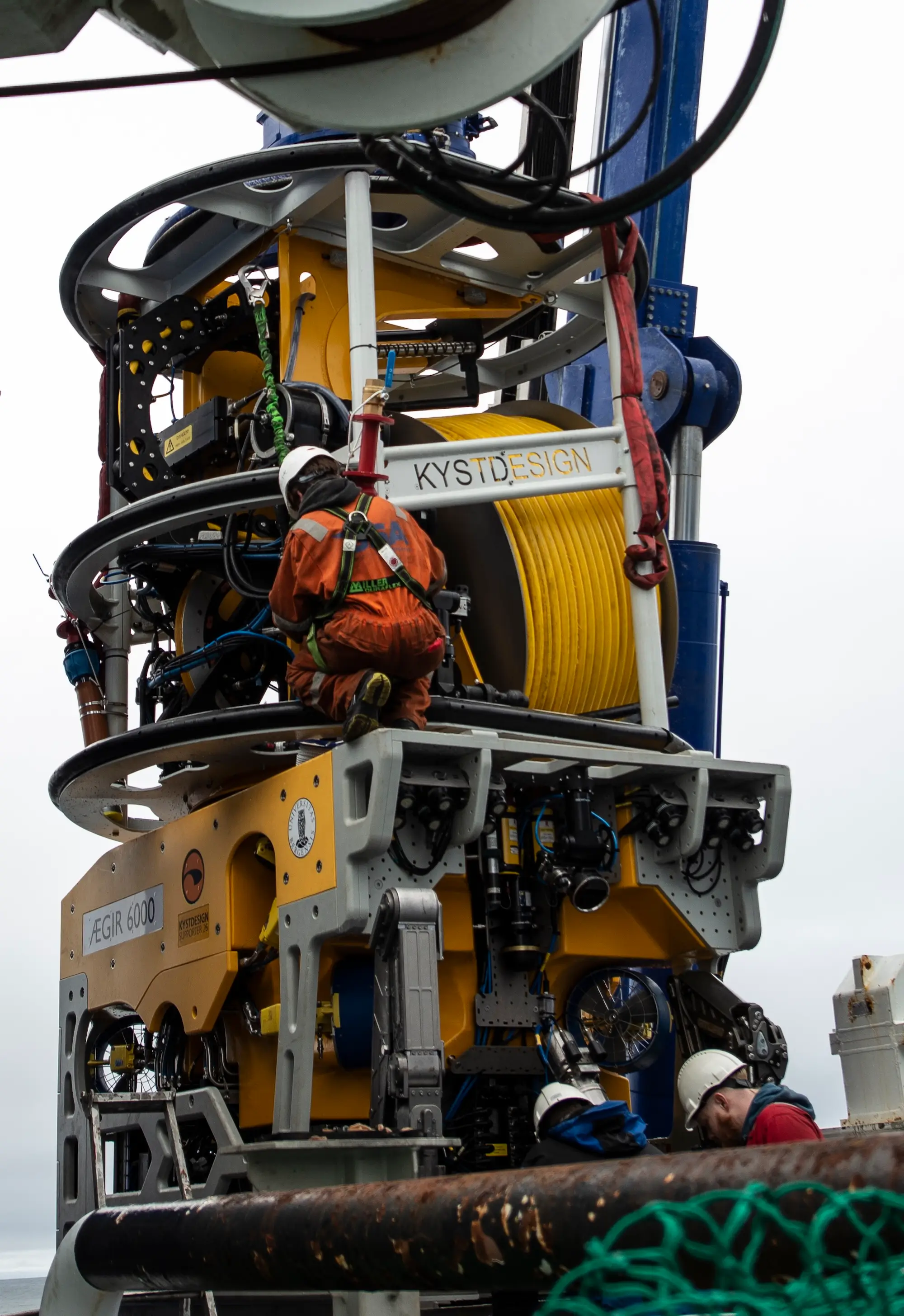 Three workers with helmets and harnesses prepare a yellow underwater robotic vehicle.