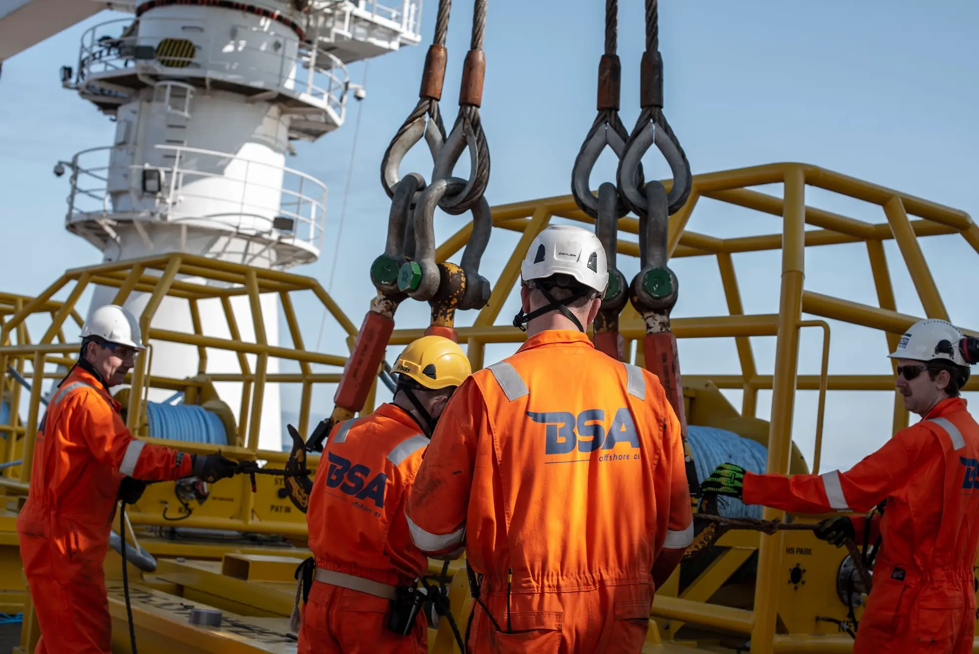 Workers in orange BSA offshore suits and helmets handling heavy lifting hooks on a yellow platform.