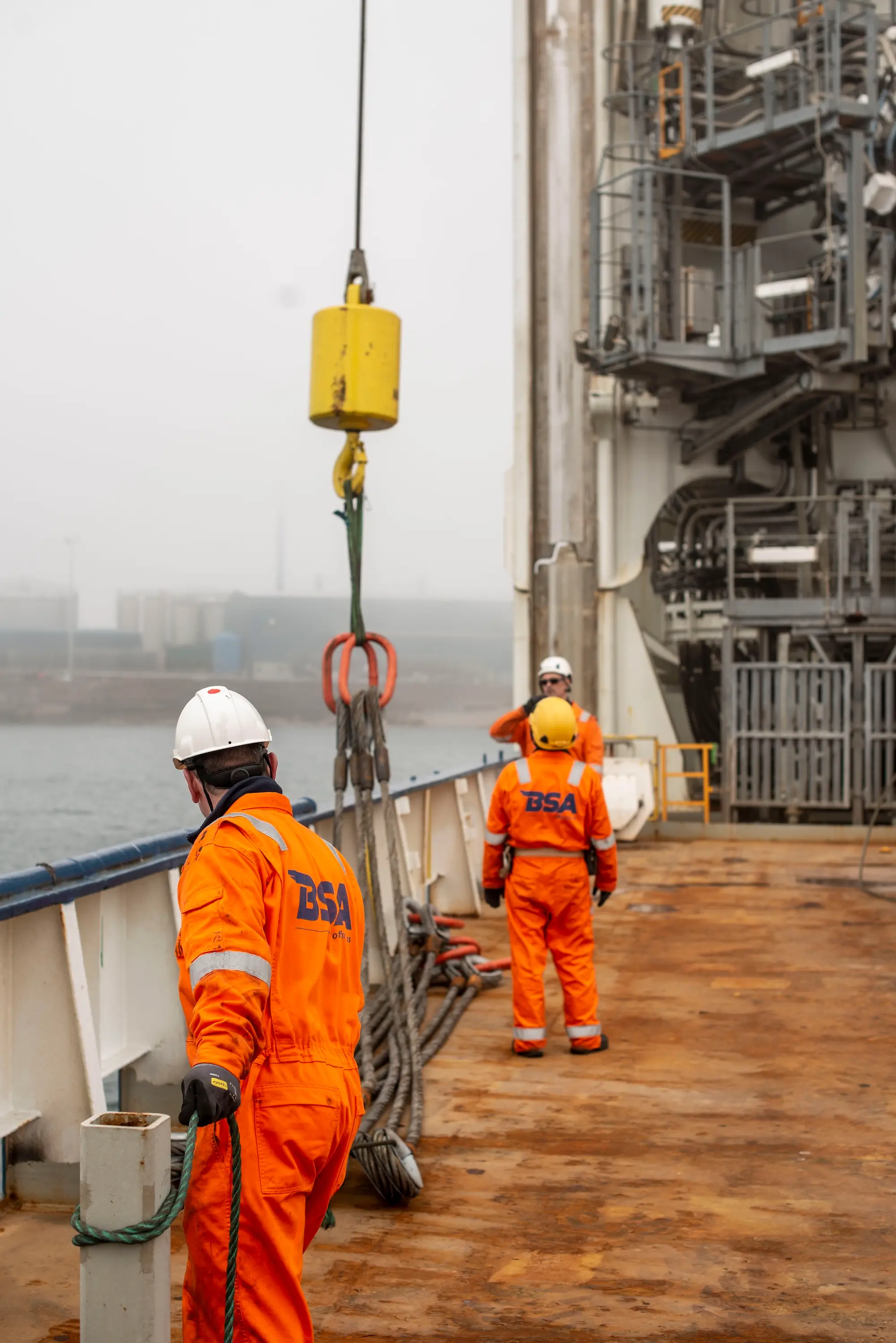 Three workers in orange BSA uniforms and helmets handling rigging on a rusty ship deck by the water.