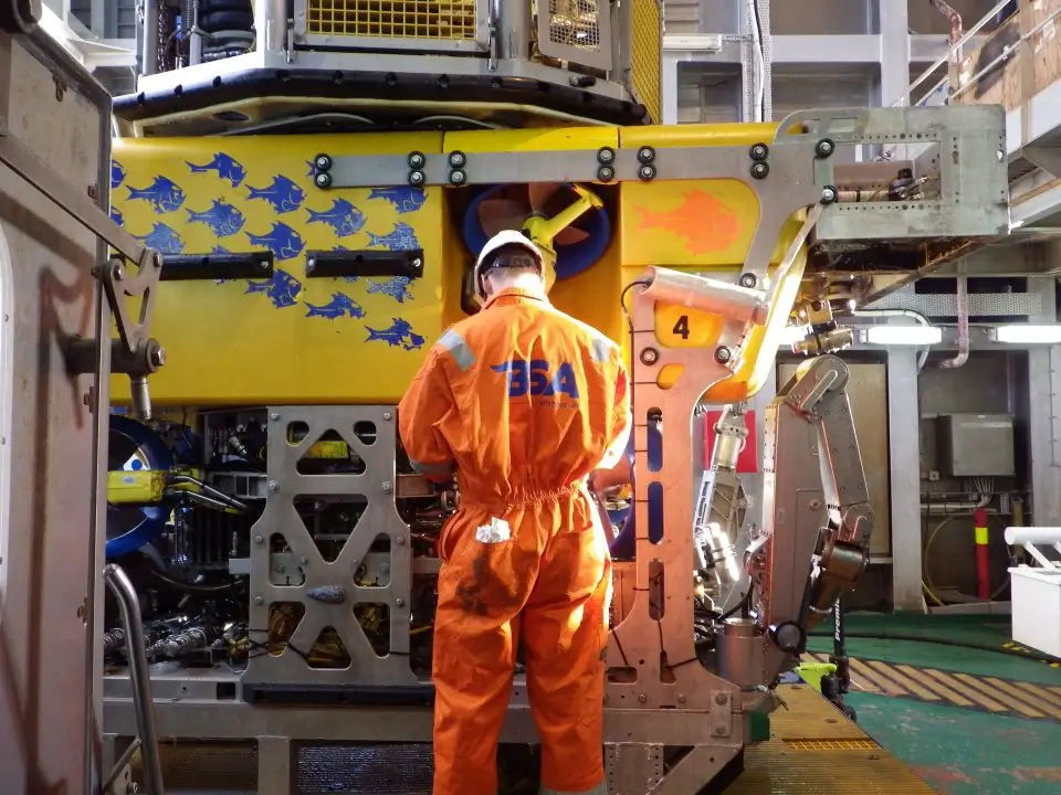 Worker in an orange suit and white helmet stands facing a yellow underwater vehicle.