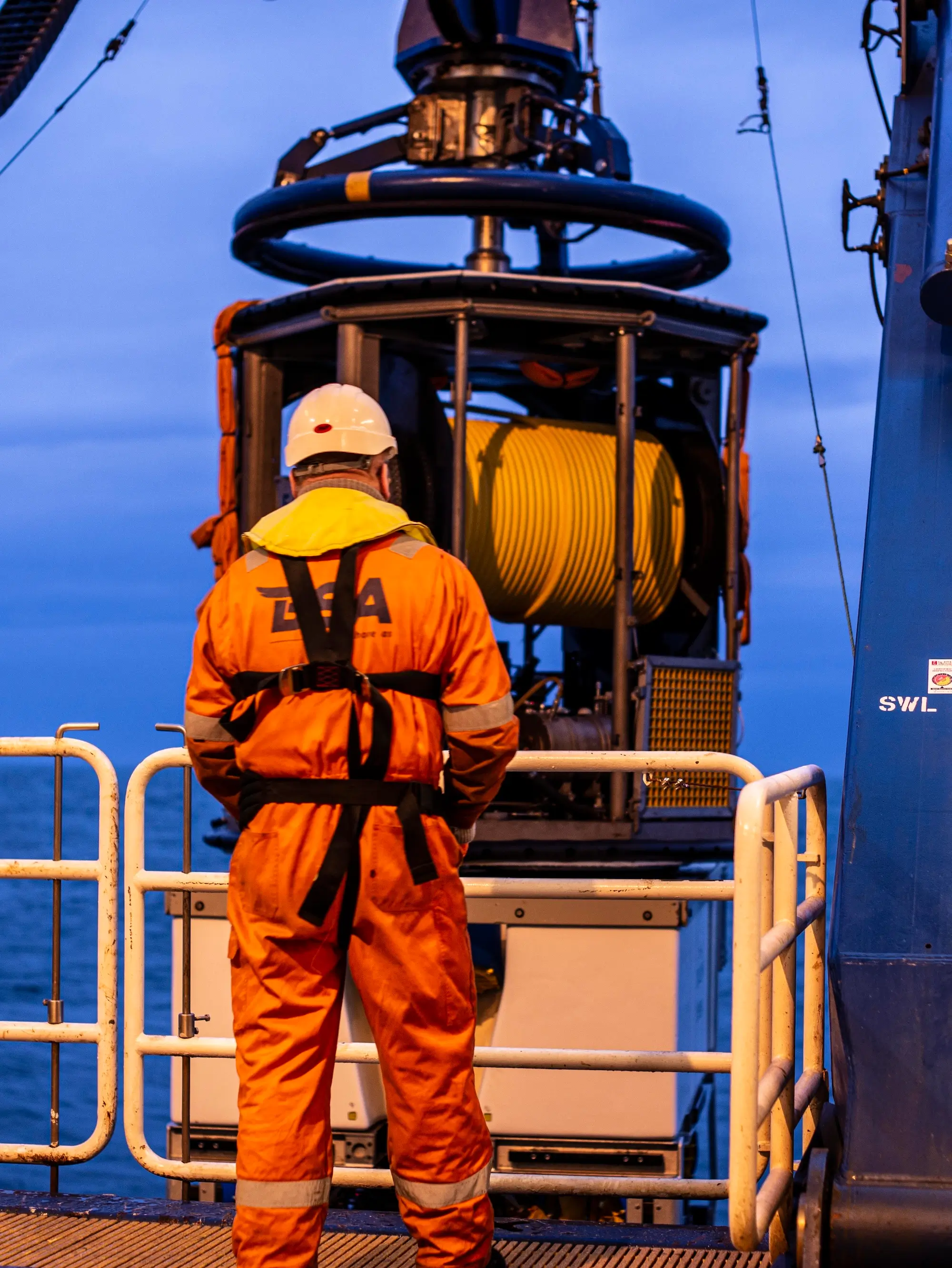 Worker in orange safety suit and helmet standing on offshore platform near large yellow cable reel.