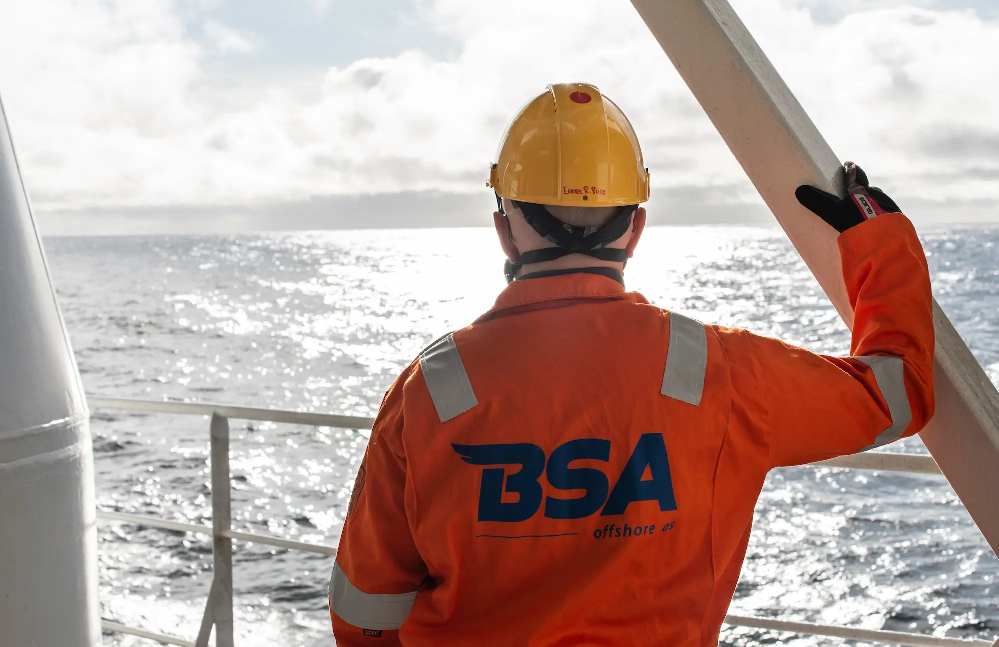 Worker in an orange jacket and yellow helmet on an offshore platform looking at the sea.