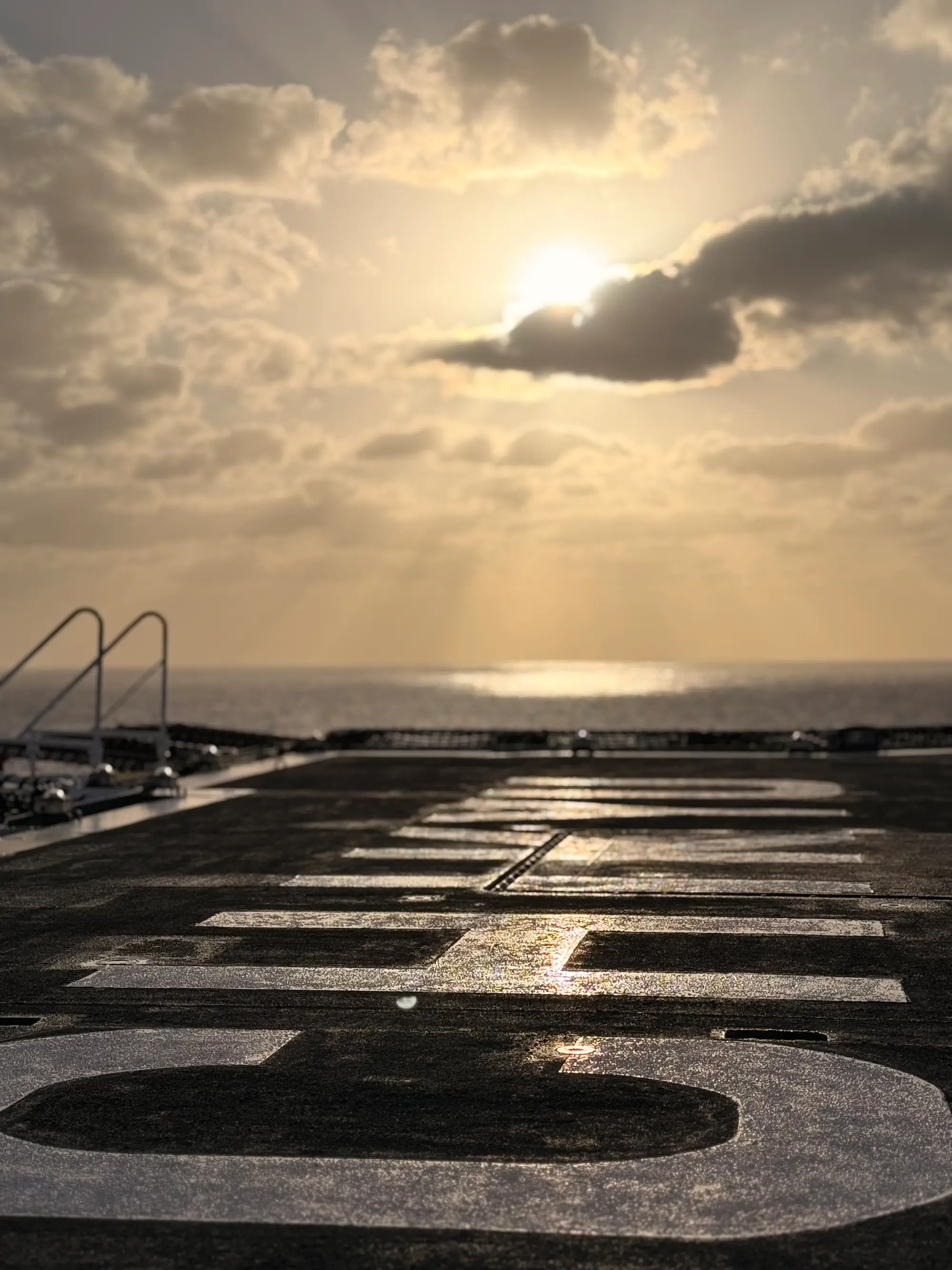 Helipad surface with painted letters facing a cloudy sky at sunrise or sunset over the sea.