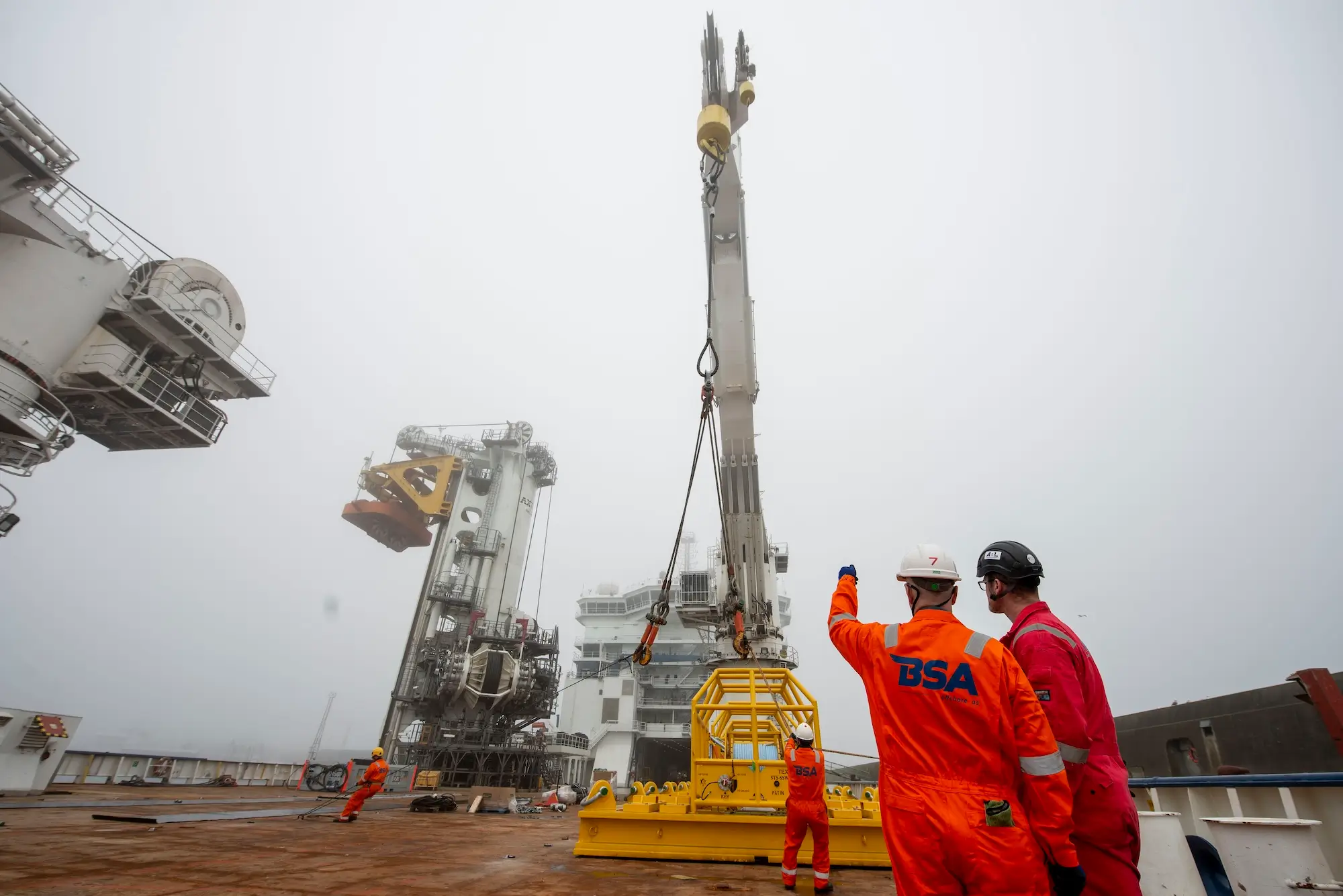 Workers in safety gear guide a large yellow structure being lifted by a crane on an offshore platform.