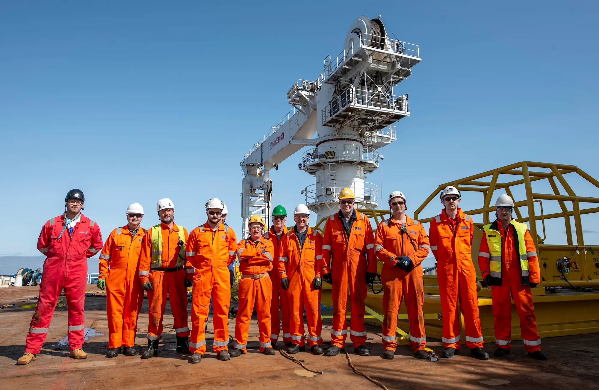 Group of offshore workers in orange safety suits and helmets standing on a platform with equipment behind.