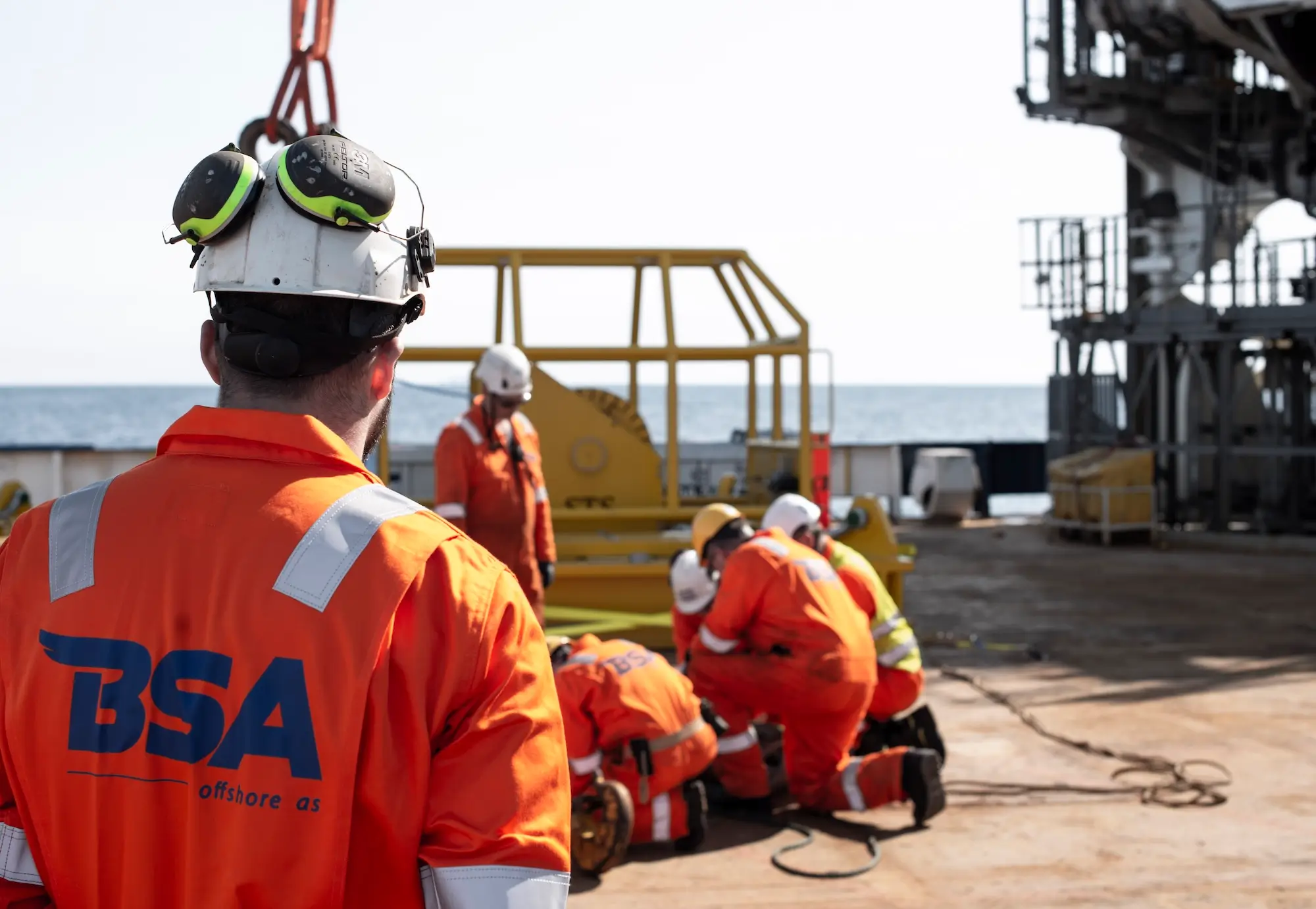 Offshore workers in orange safety gear and helmets working on a platform at sea under clear sky.