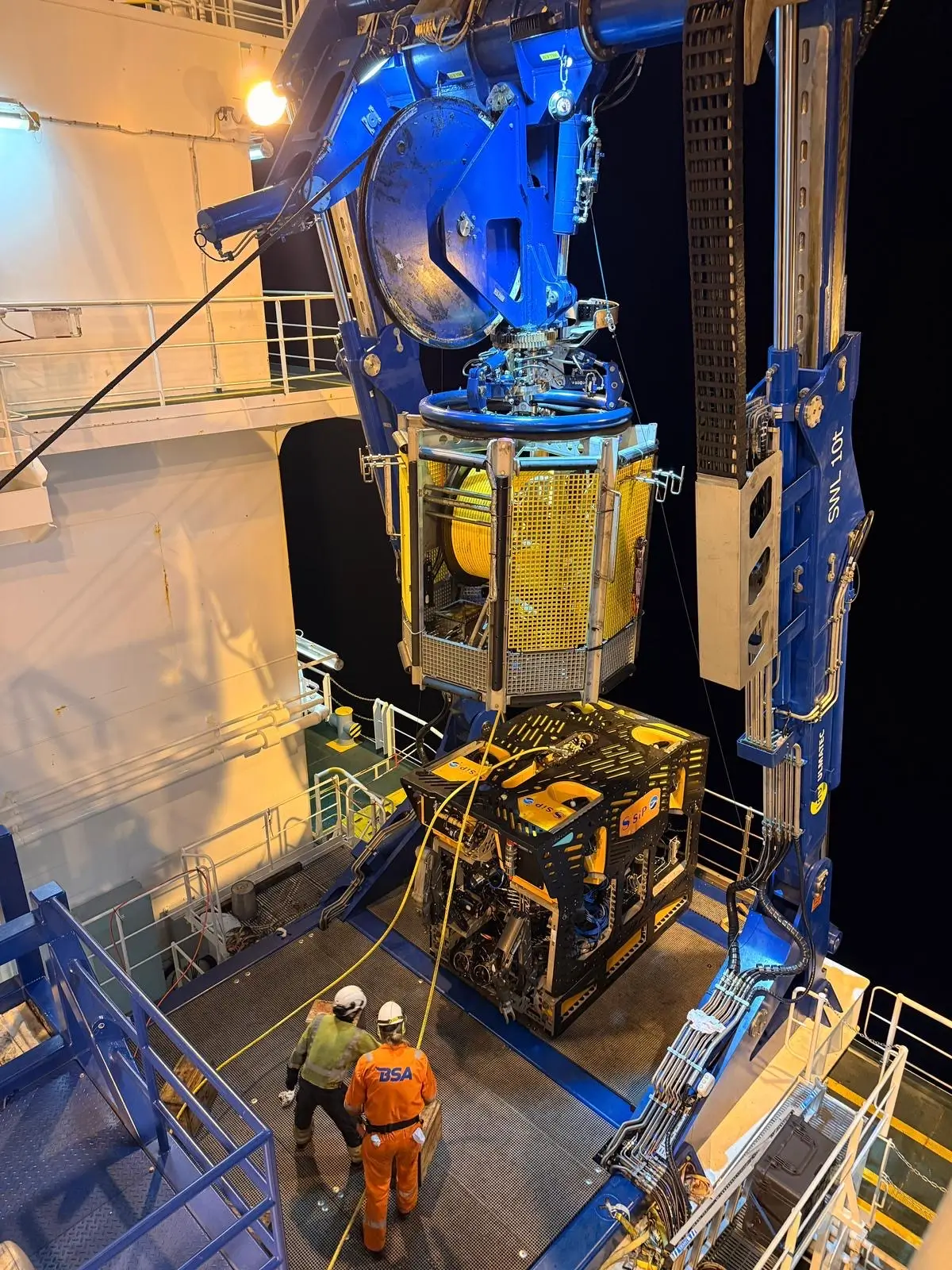 Two workers handling underwater ROV being lowered by ship crane at night.