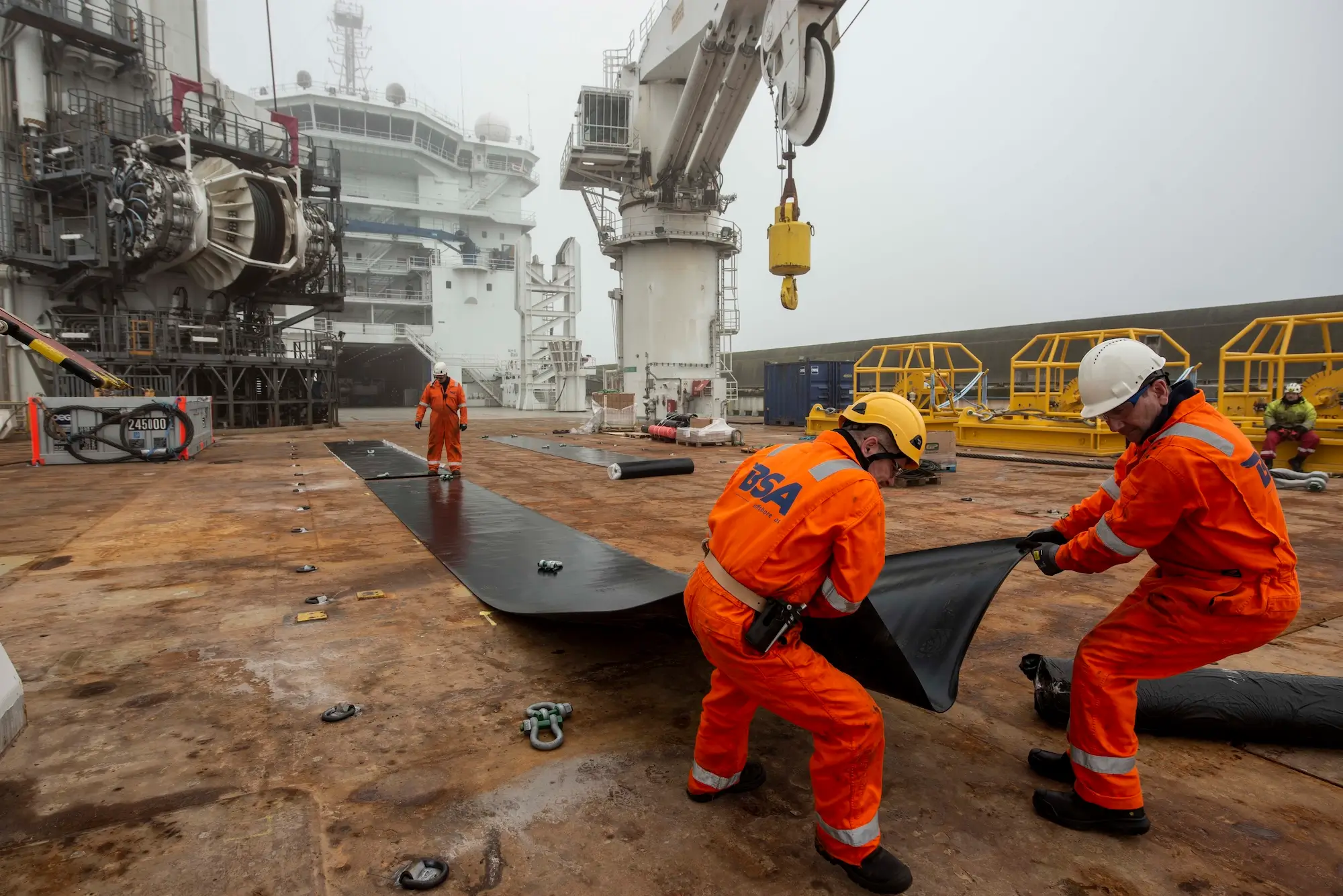 Workers in orange safety gear and helmets unfurl a large black mat on an industrial ship deck.