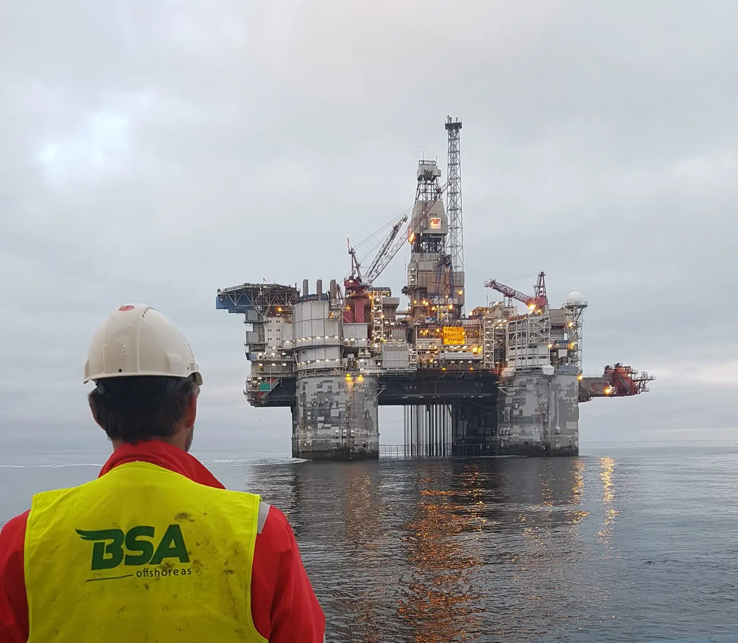 Worker in safety gear faces a lit offshore oil platform on calm water under a cloudy sky.