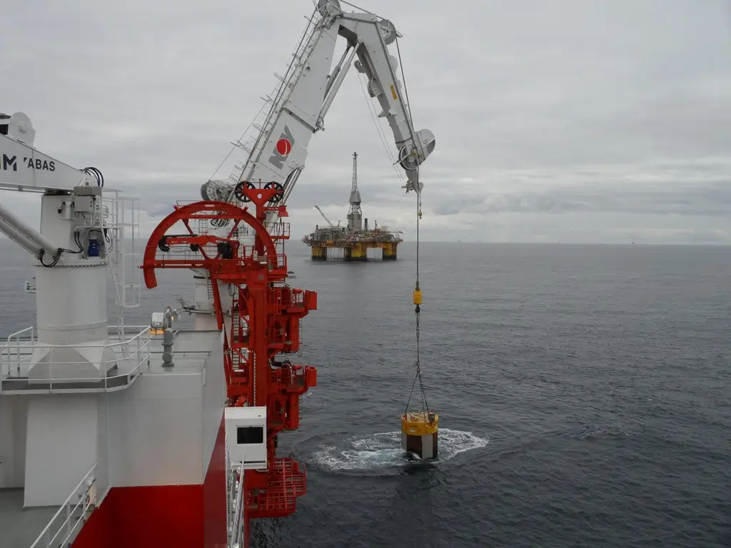 Crane on a ship lowering equipment into the sea with an offshore oil platform in the background.