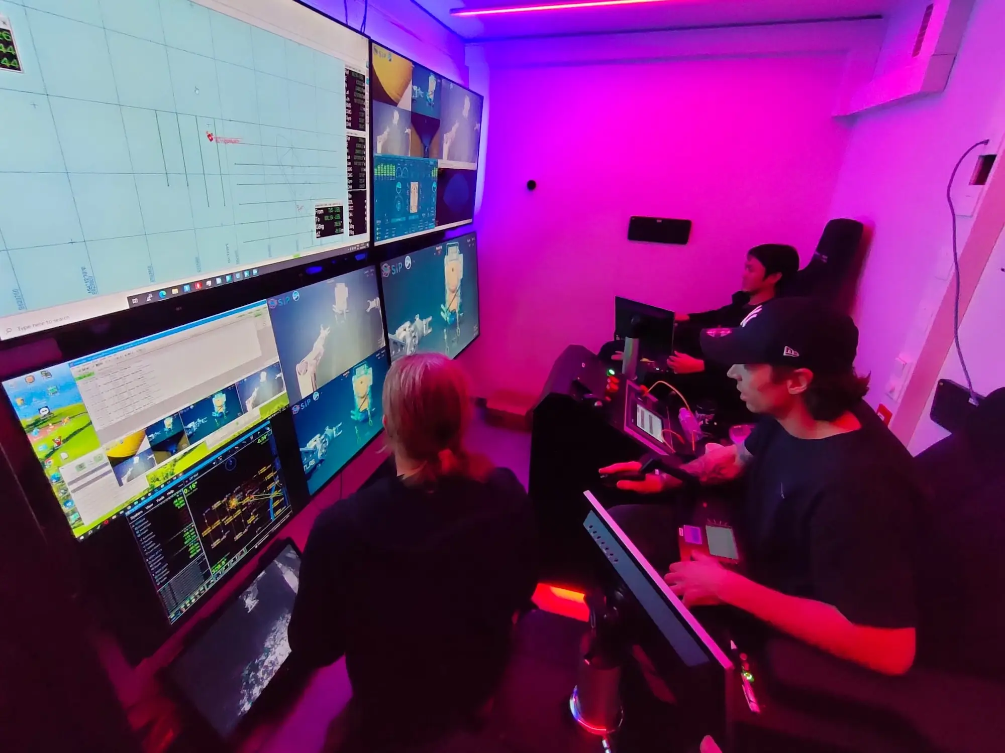 Two people operate multiple computer screens with marine robotics and map data in a purple-lit control room.