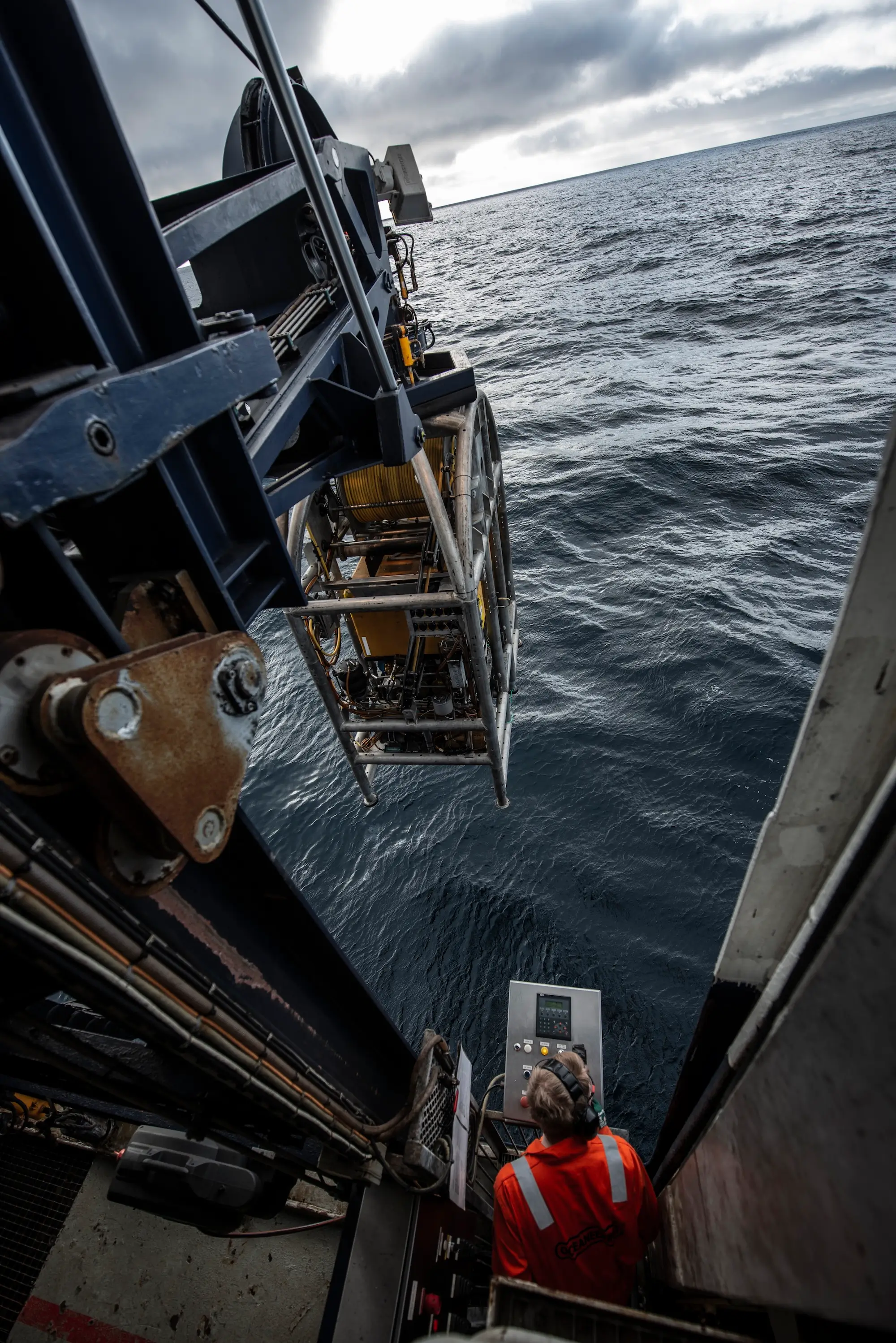 Worker in orange gear operating machinery on a ship deploying equipment into the ocean under cloudy skies.