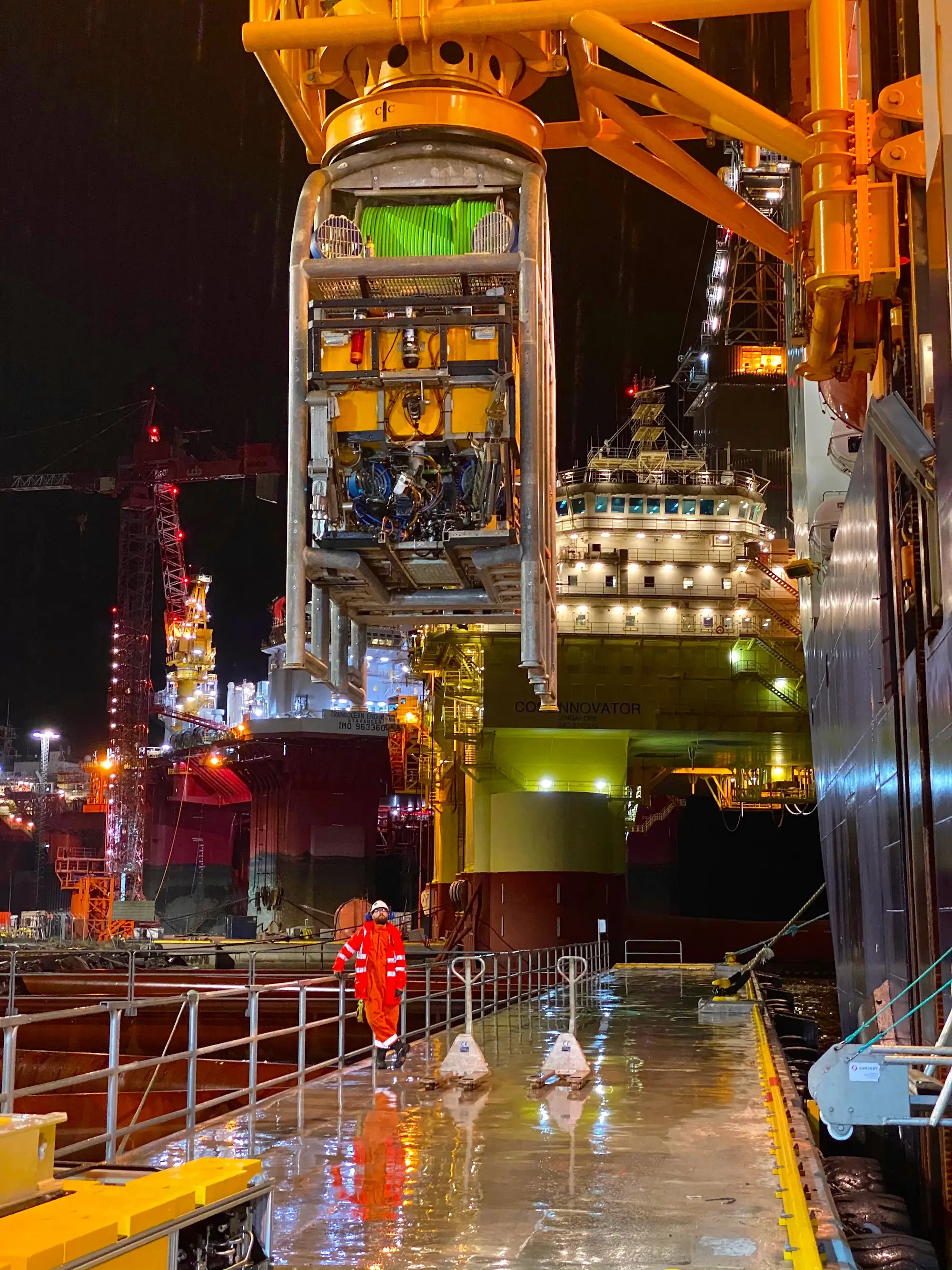 Worker in orange gear walking on wet platform near large offshore oil rig with machinery overhead at night.