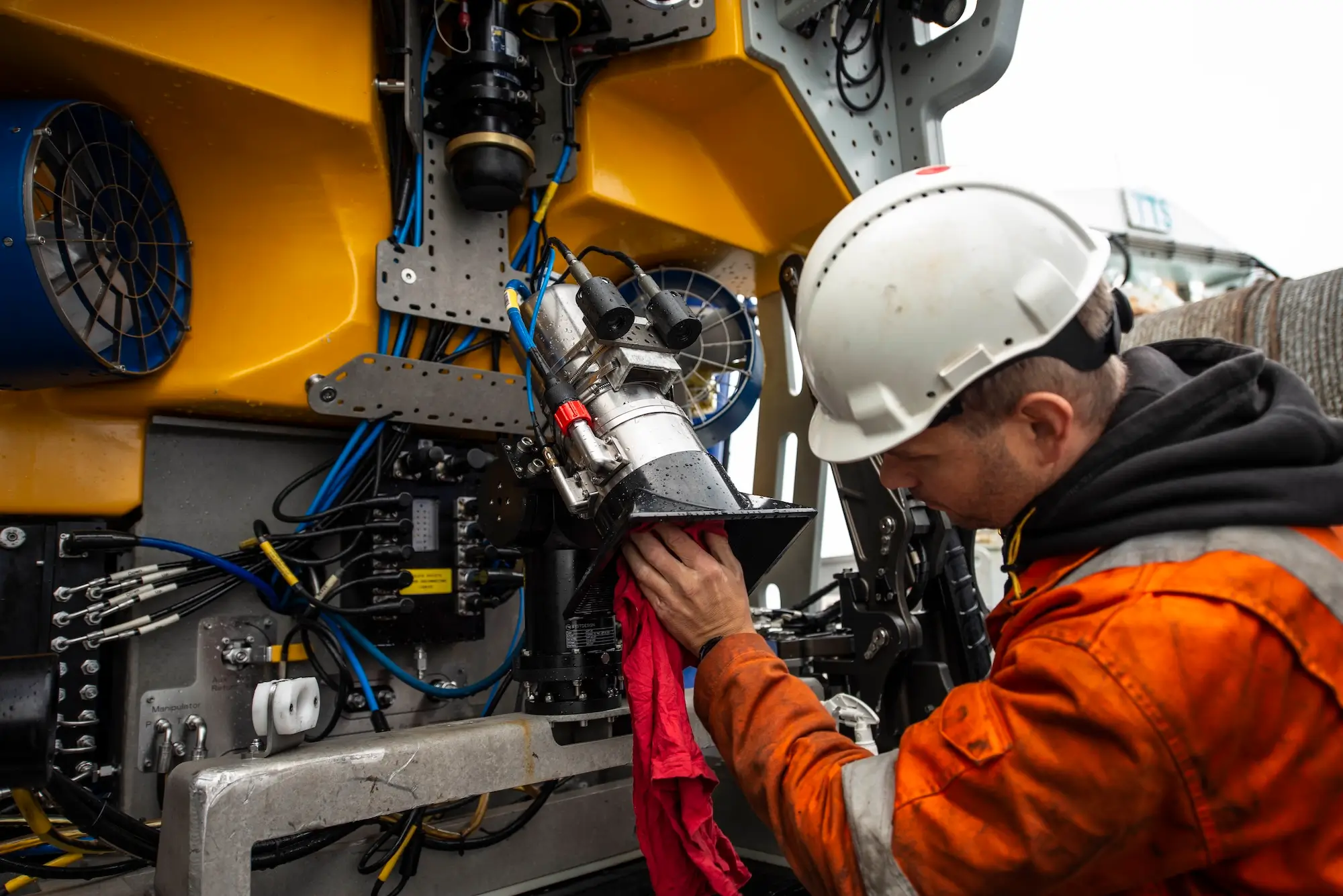 Worker in helmet and orange safety gear cleaning a machine part with a red cloth.