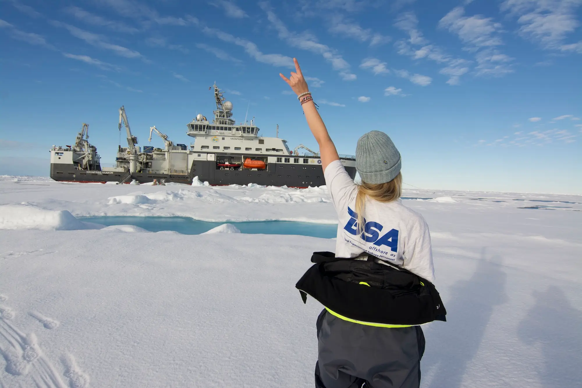 Person in a BSA Offshore shirt and beanie raises hand making a rock sign near an ice-covered sea and ship.
