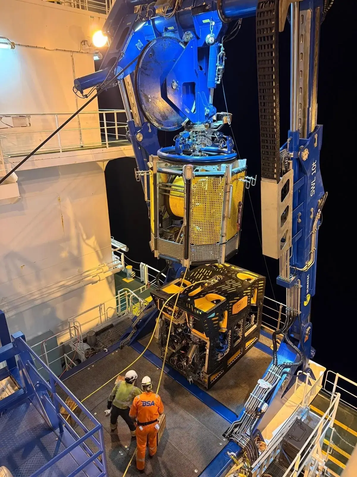 Two workers secure an remotely operated underwater vehicle on a vessel using a crane at night.