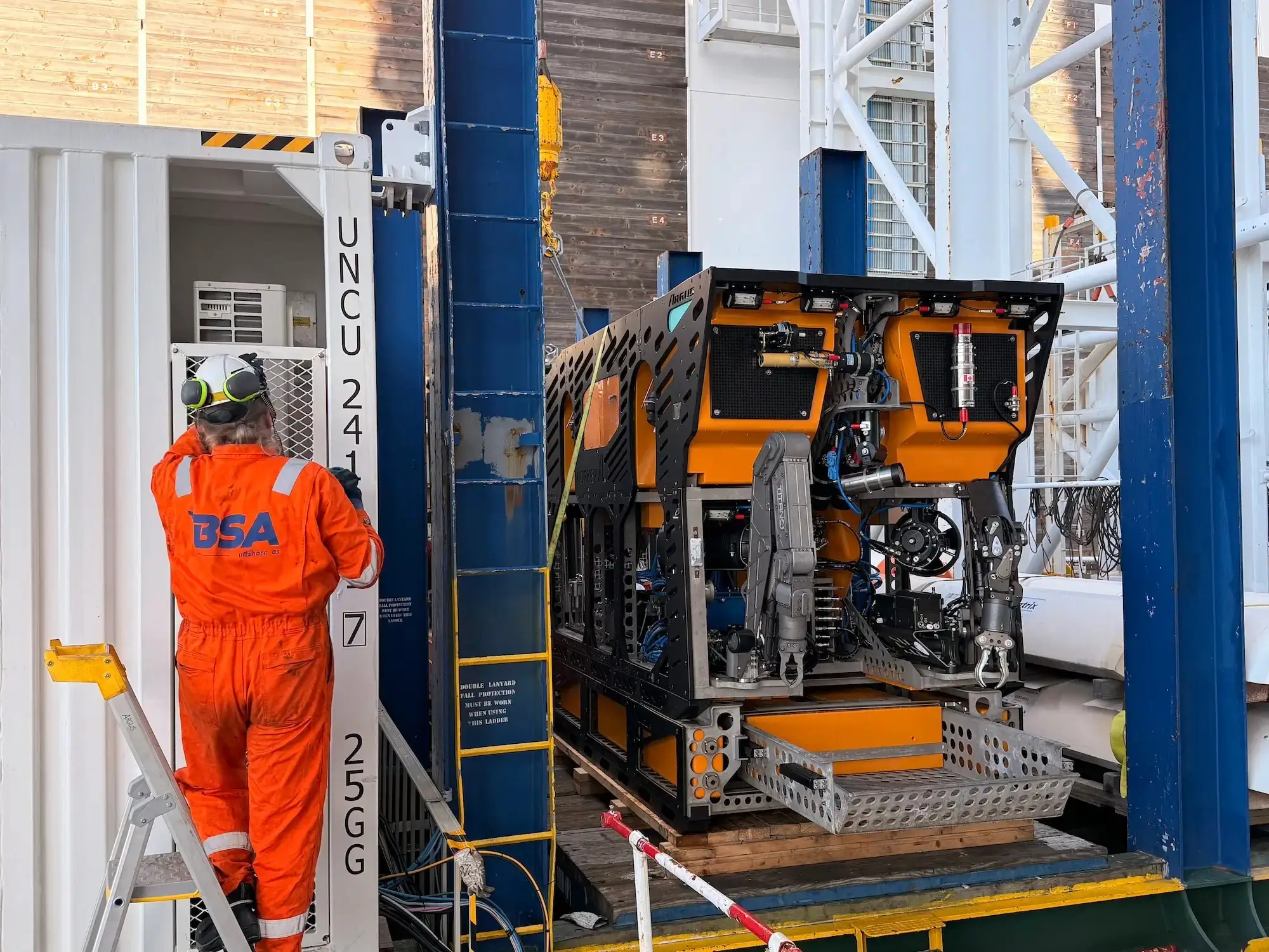 Worker in orange BSA suit standing near a robotic underwater vehicle on a ship deck.