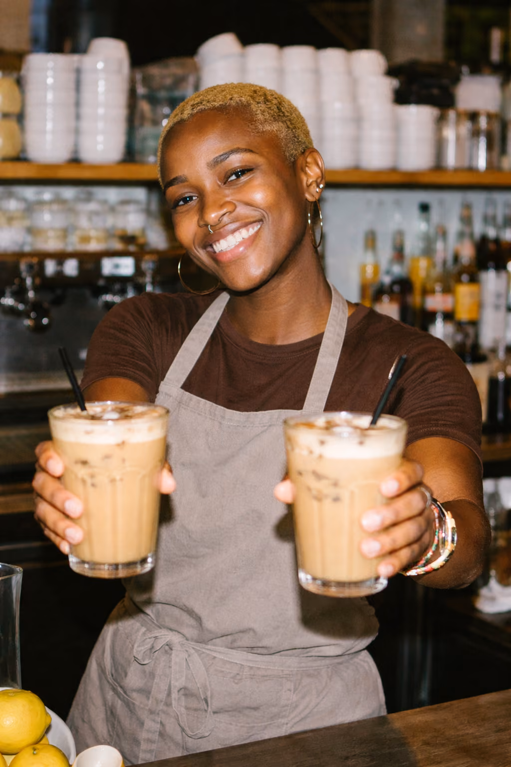 Smiling barista in apron holding two iced coffee drinks with straws at a café counter.