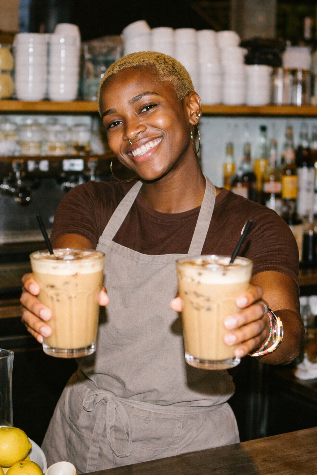 Smiling barista in apron holding two iced coffee drinks with straws at a café counter.