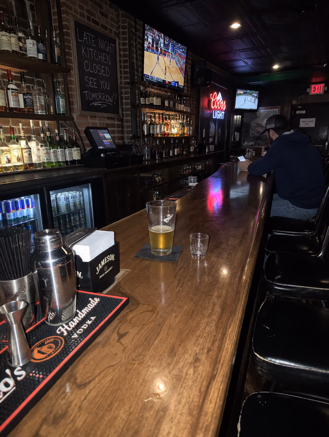 Man in cap and hoodie sits at dimly lit bar counter with drinks, TVs showing basketball and hockey games.