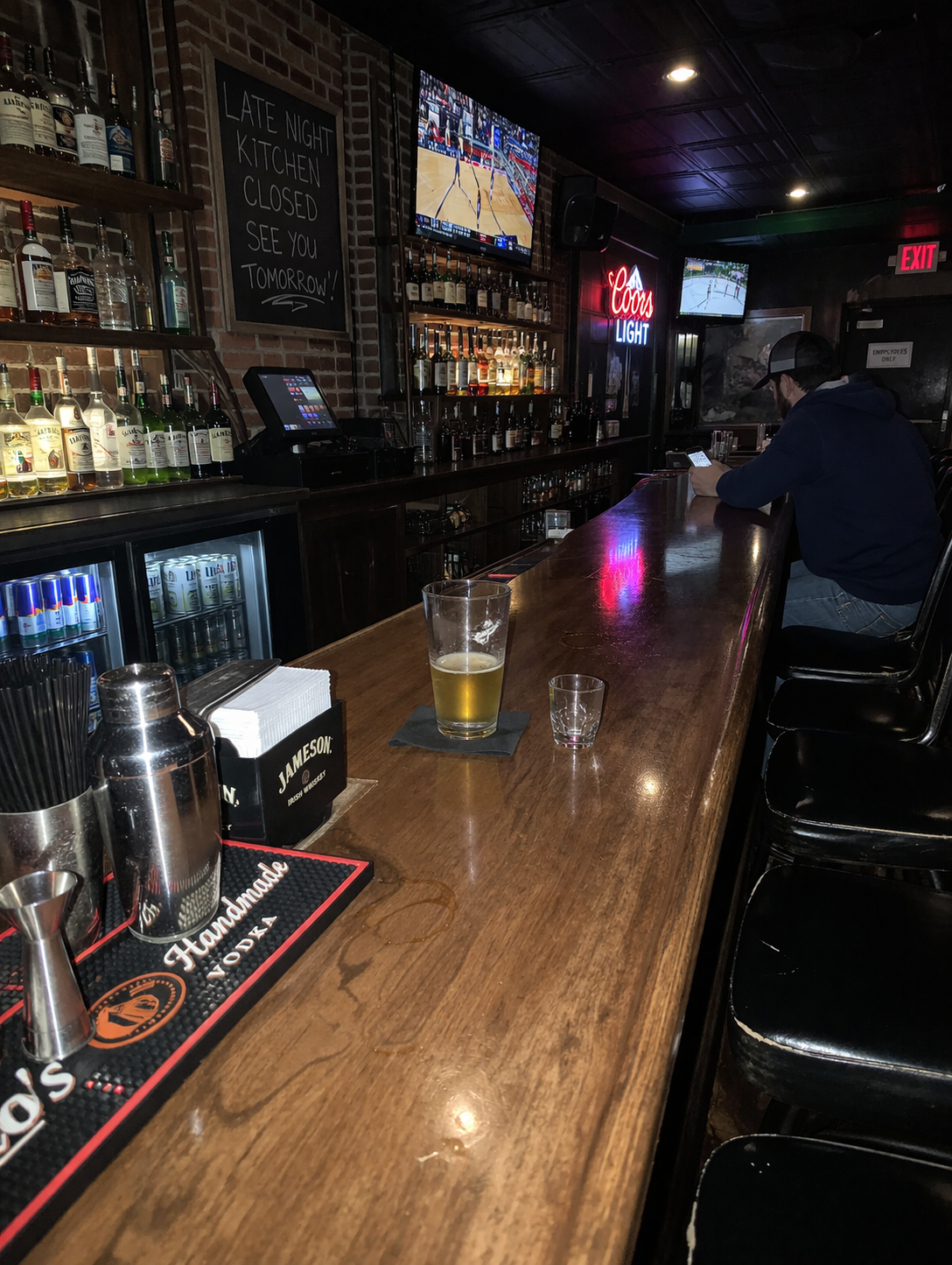 Man in cap and hoodie sits at dimly lit bar counter with drinks, TVs showing basketball and hockey games.