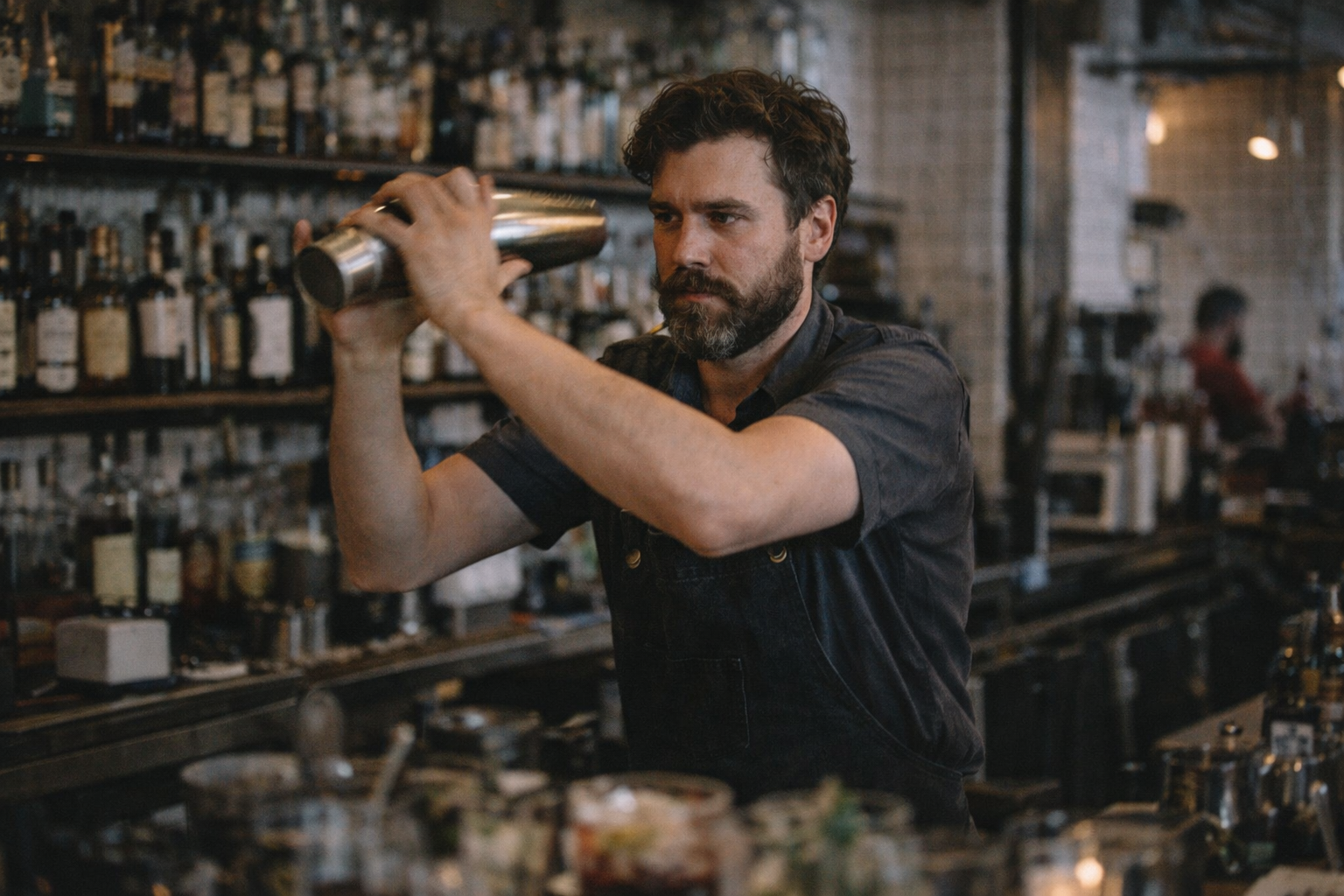 Bearded bartender shaking a cocktail shaker behind a bar with bottles on shelves in the background.