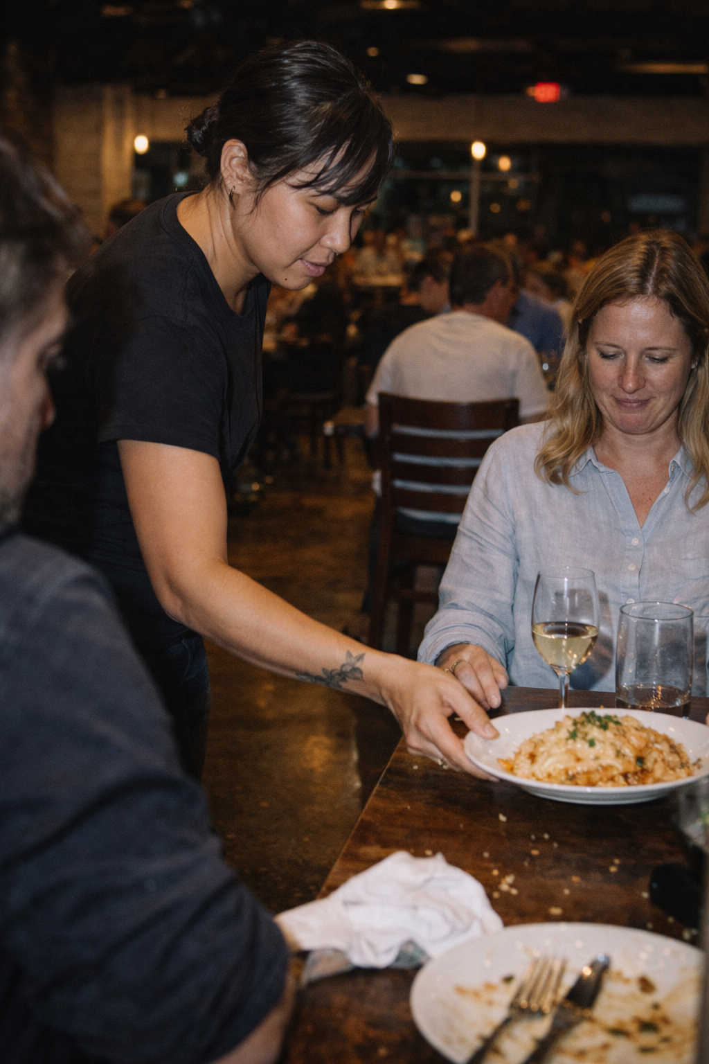 Server placing a pasta dish on a restaurant table where a woman is seated with a glass of white wine.